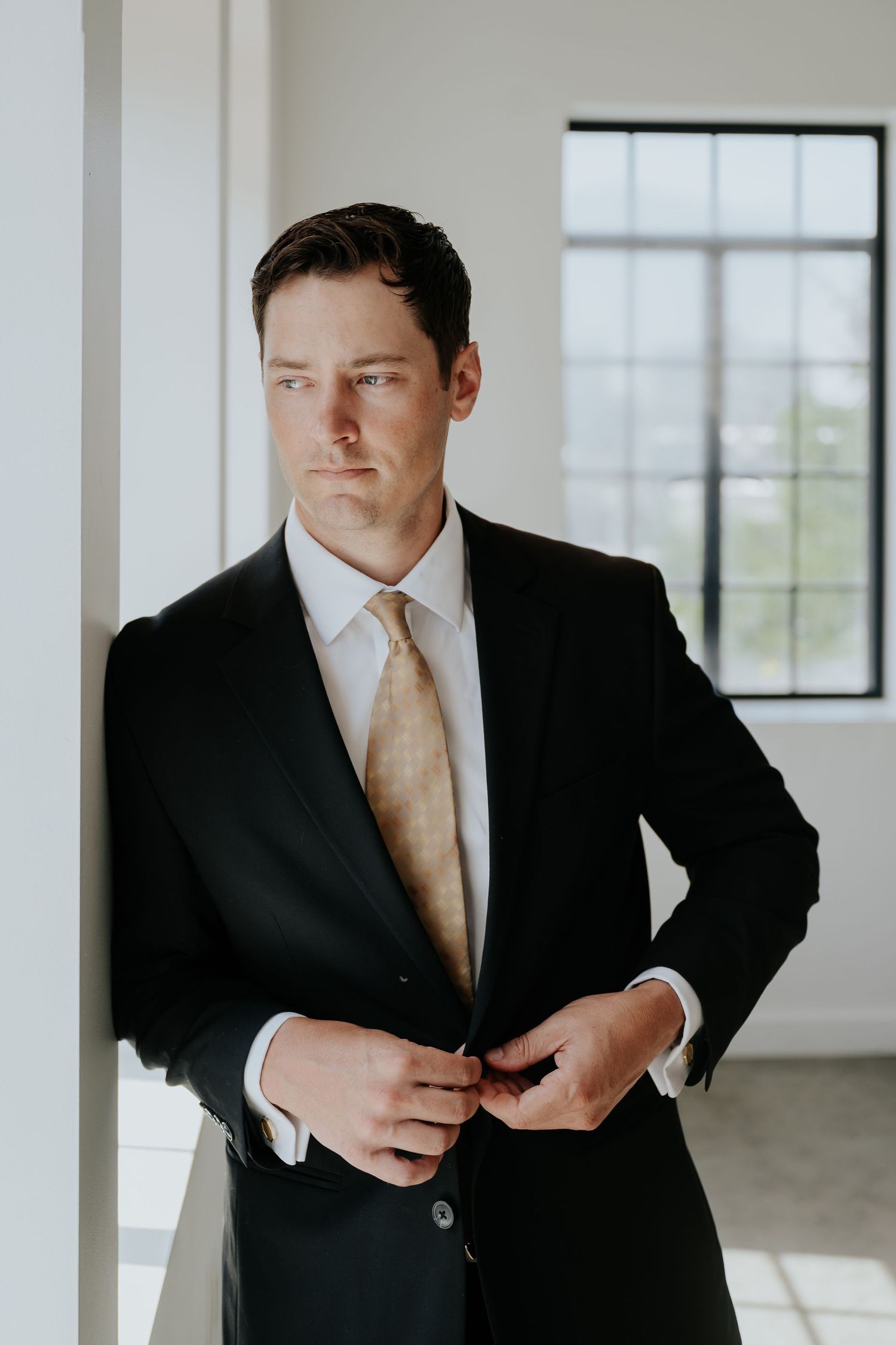Groom in black suit and gold tie buttoning jacket while leaning against white column in bright room.