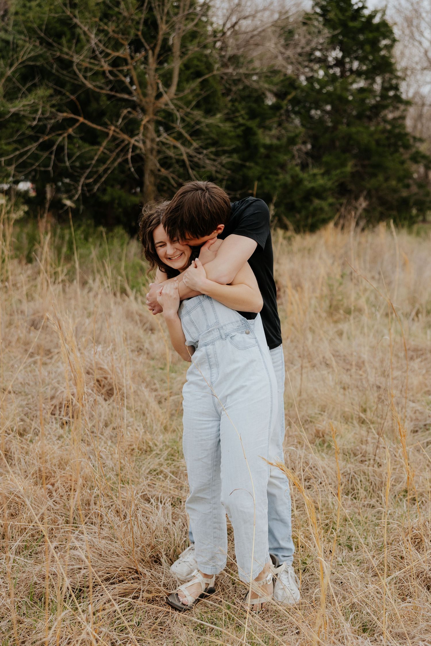 Couple embracing in a golden field, woman in light blue overalls smiling as man hugs her from behind near trees.