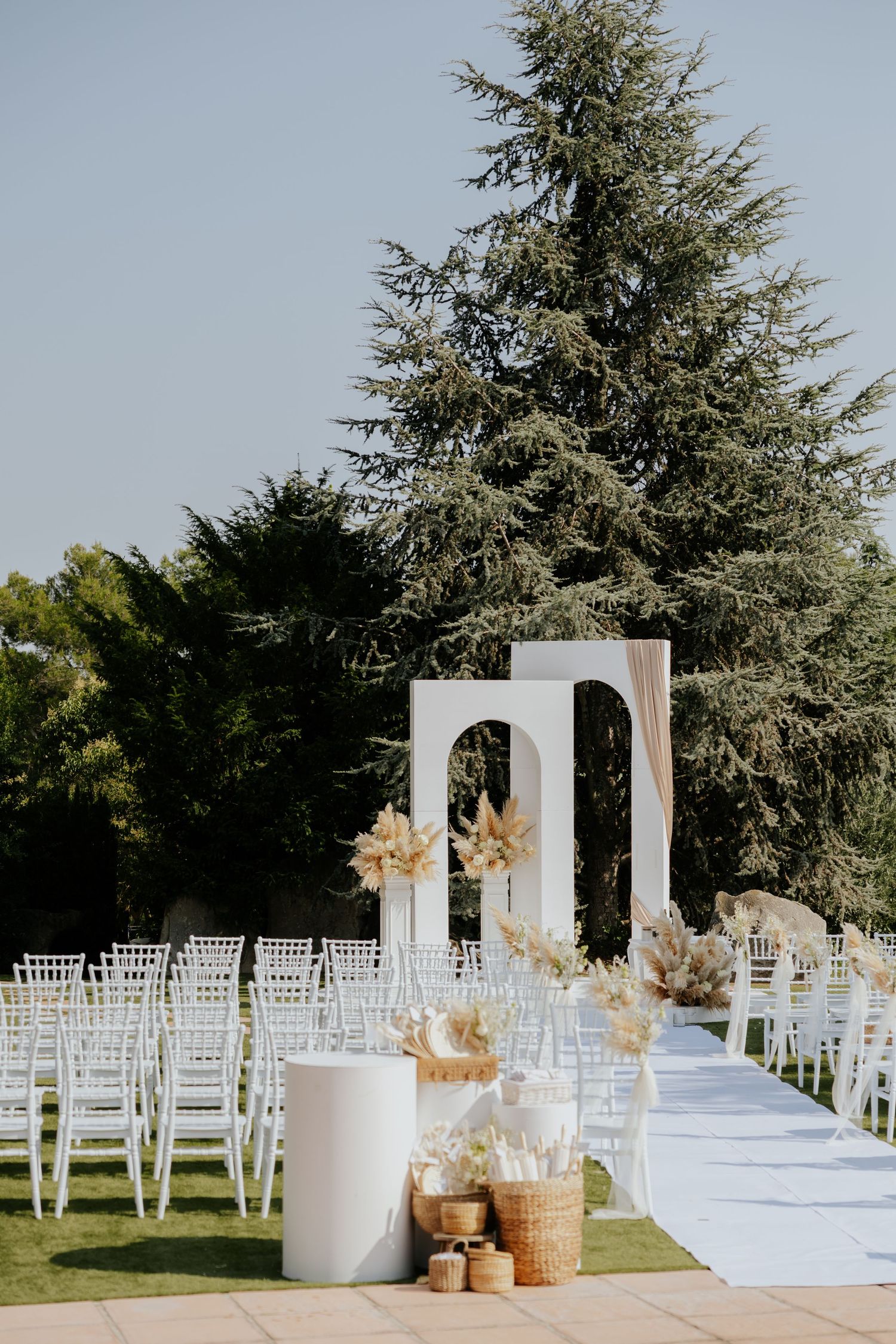 Elegant outdoor wedding ceremony setup with white chairs, arch, dried floral arrangements, and tall pine trees under clear sky.