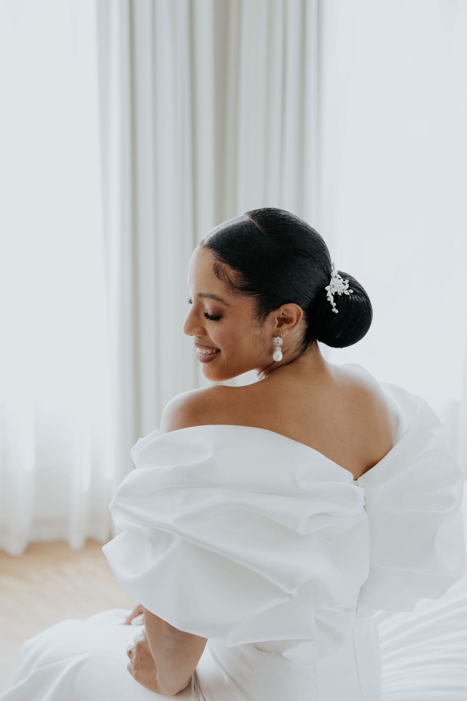 Smiling bride in white off-shoulder gown with elegant updo and pearl earrings, seated by bright window.