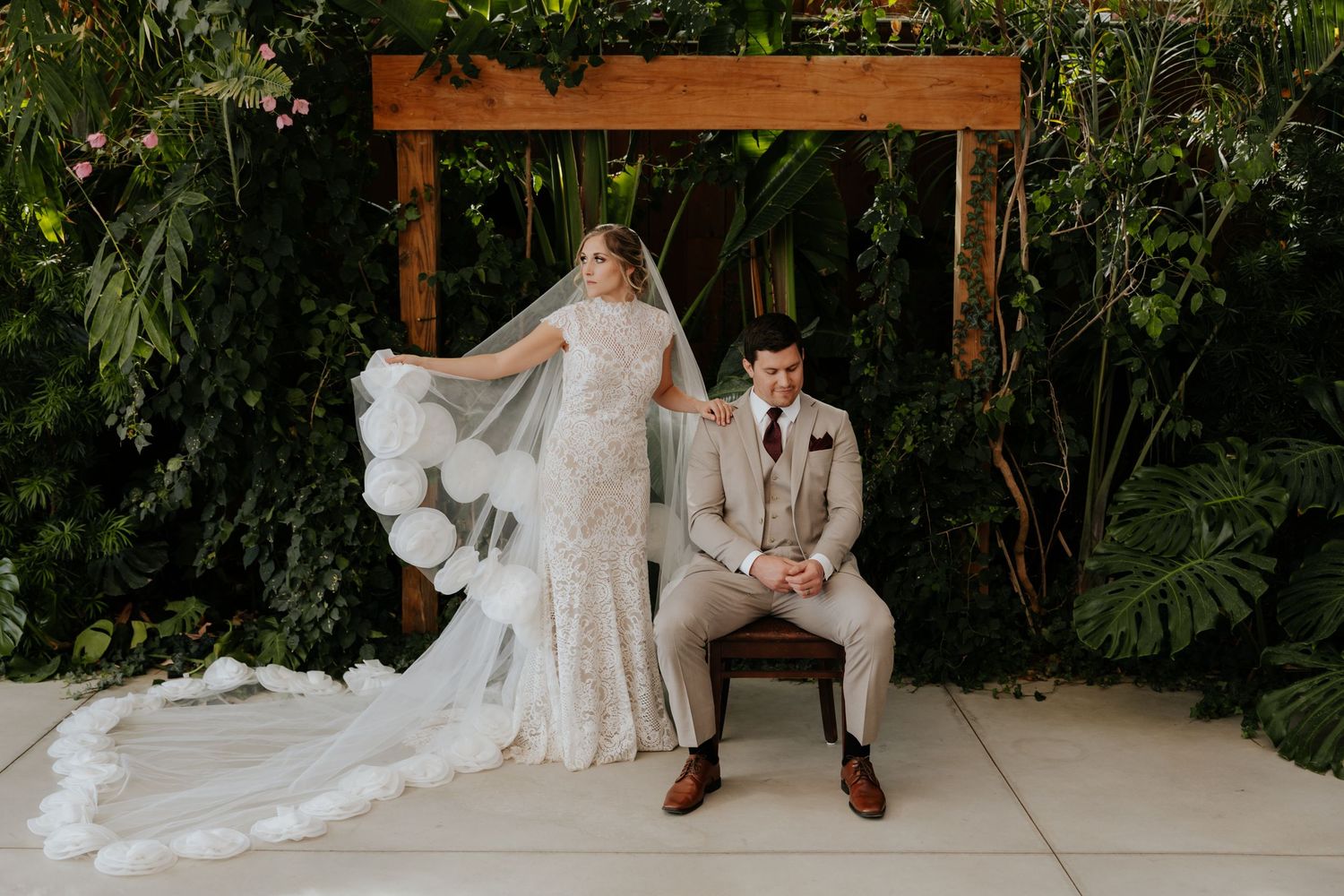 Bride in lace gown with veil stands beside groom in tan suit before lush greenery wedding backdrop.