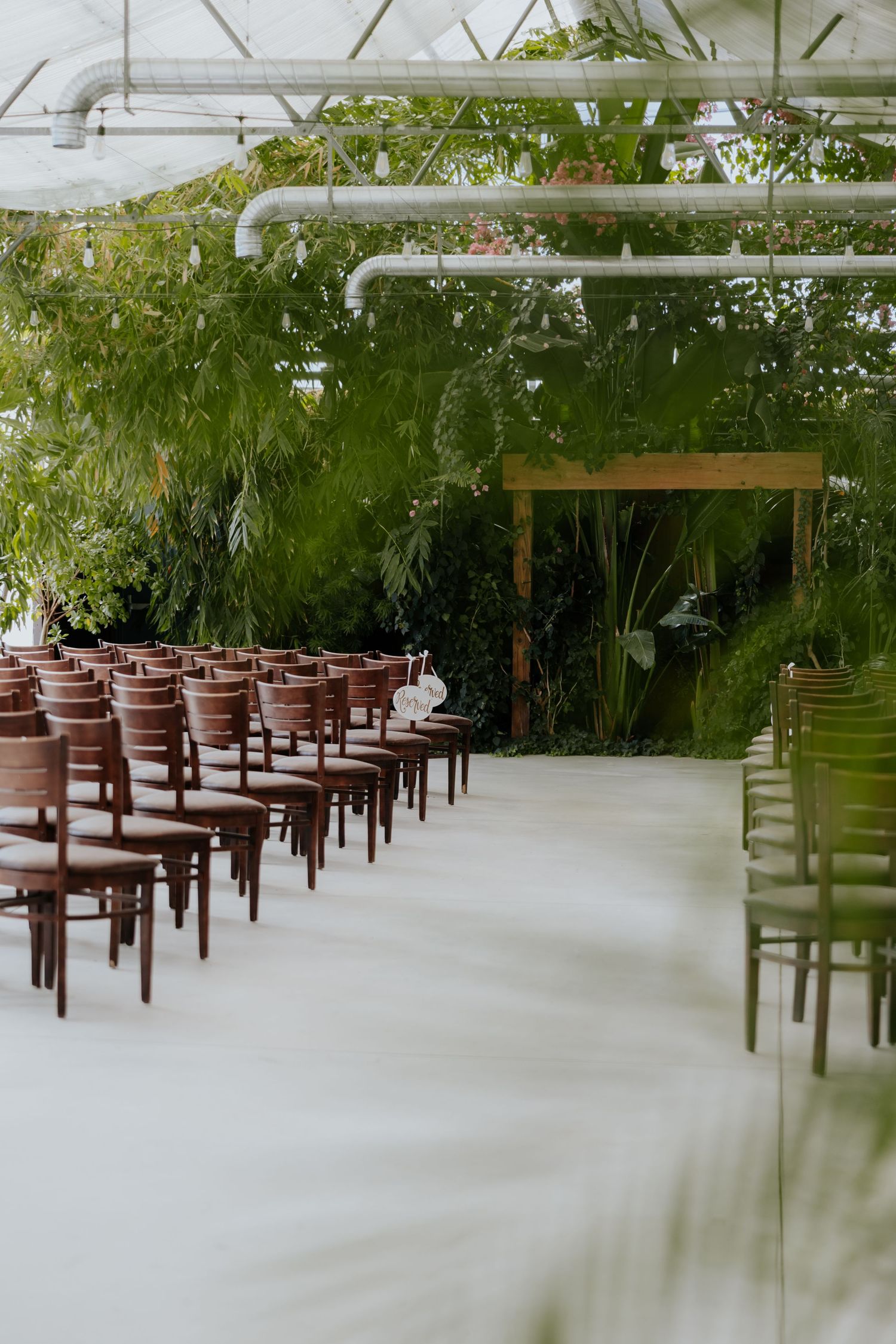 Elegant indoor wedding ceremony venue with wooden chairs, tropical greenery, and wooden arch altar.