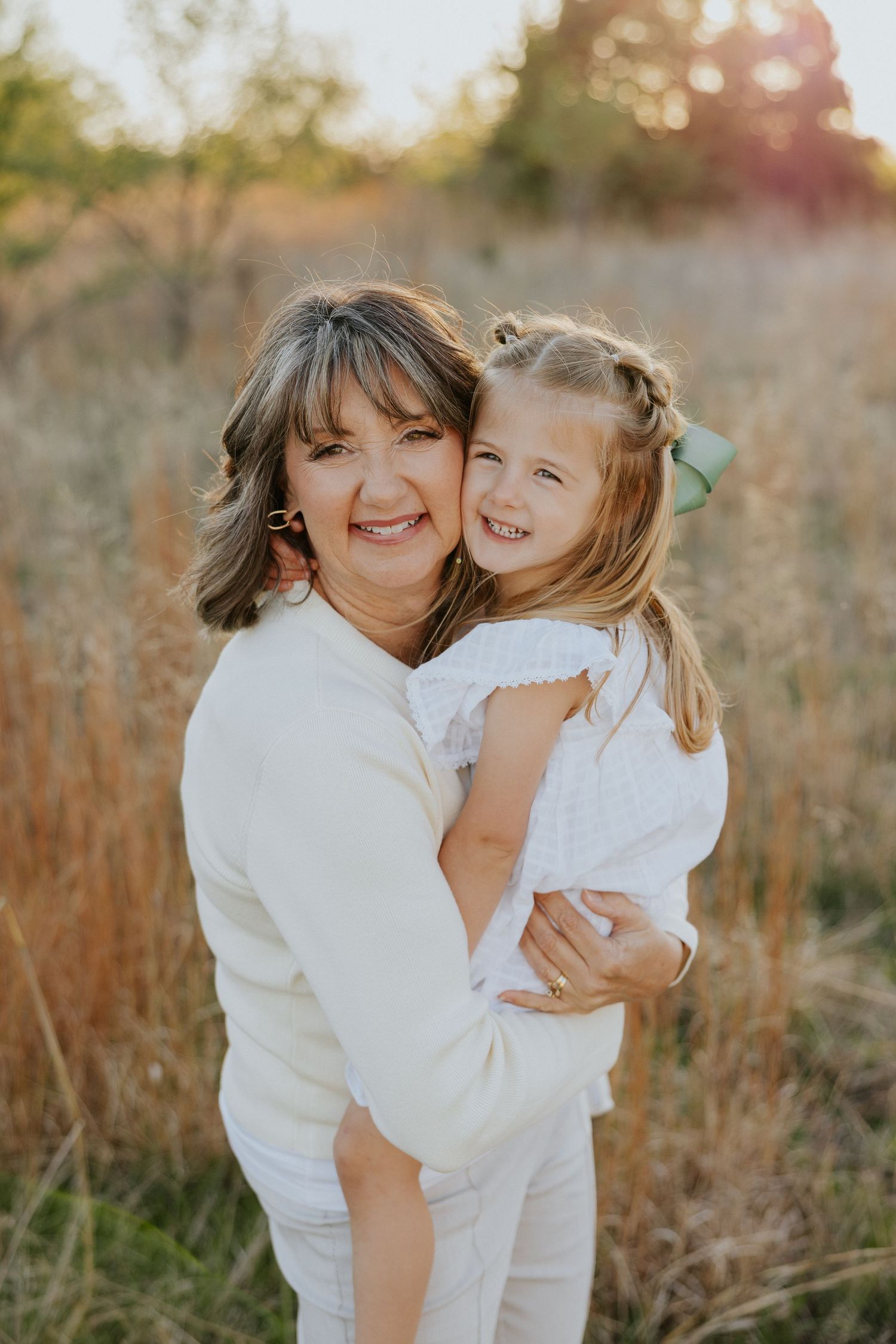 Smiling grandmother hugs her young granddaughter in a sunlit field during a family photo session.