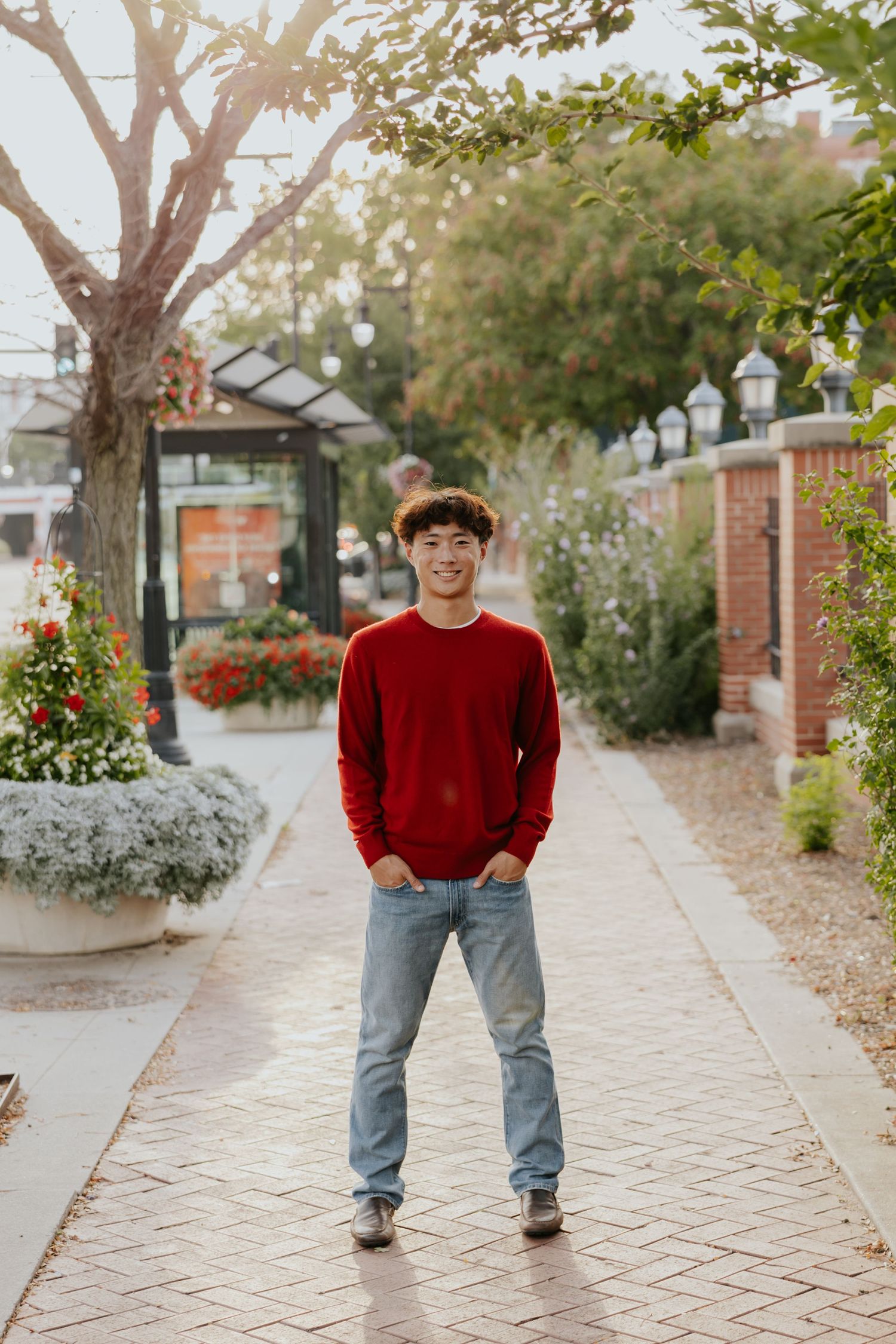 Smiling young man in a red sweater and jeans stands on a brick walkway surrounded by flowers.