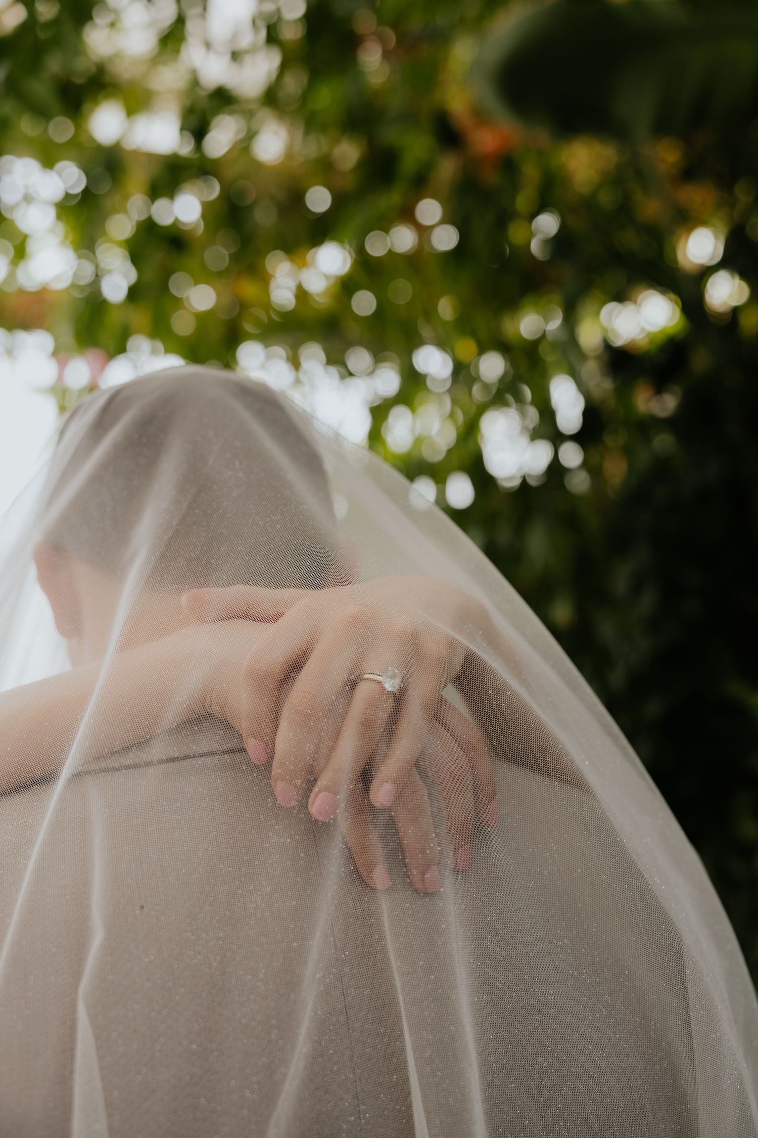 Bride resting hand with diamond engagement ring on grooms shoulder beneath flowing white veil.