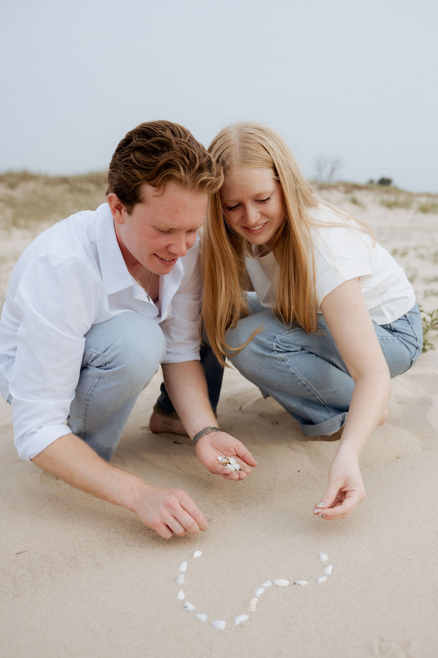 Couple crouching on sandy beach arranging seashells into a heart shape together.