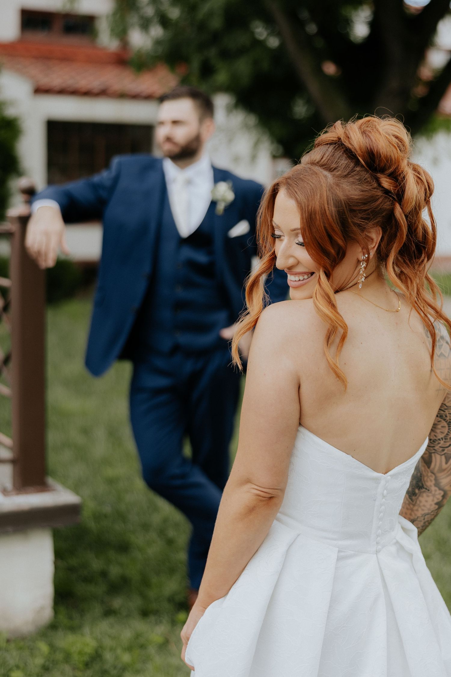 Smiling red-haired bride in strapless gown glancing back at groom in navy suit outdoors.