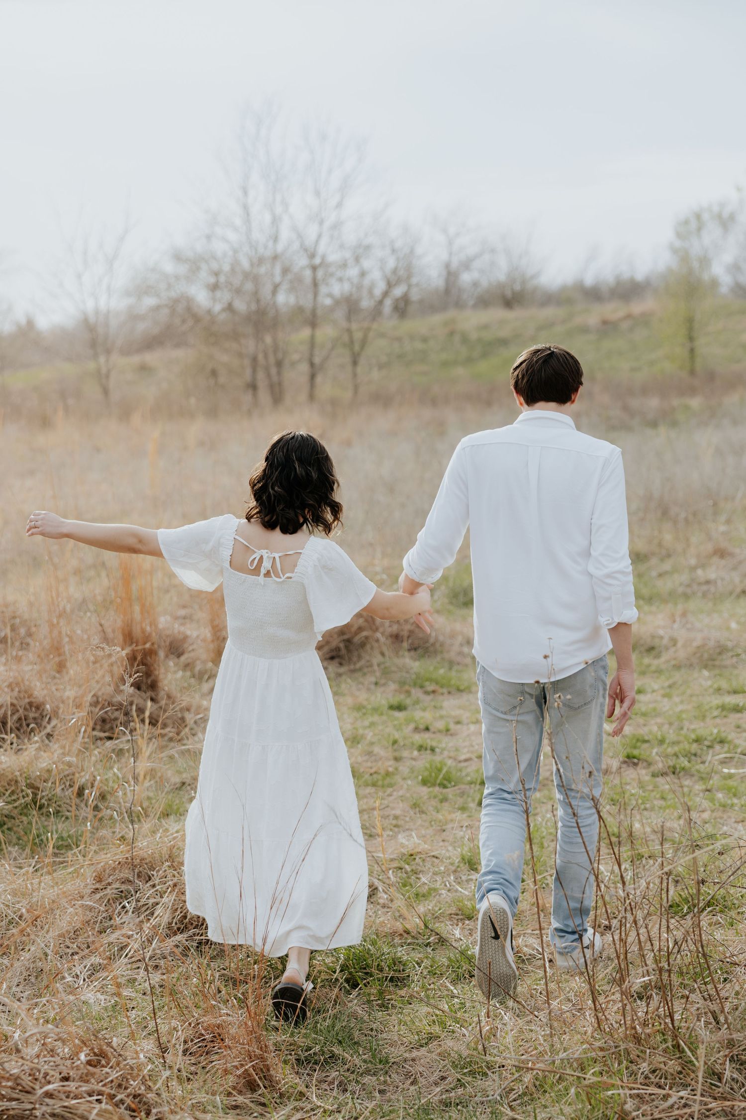 Couple in white outfits walking hand in hand through an open field during engagement photos.
