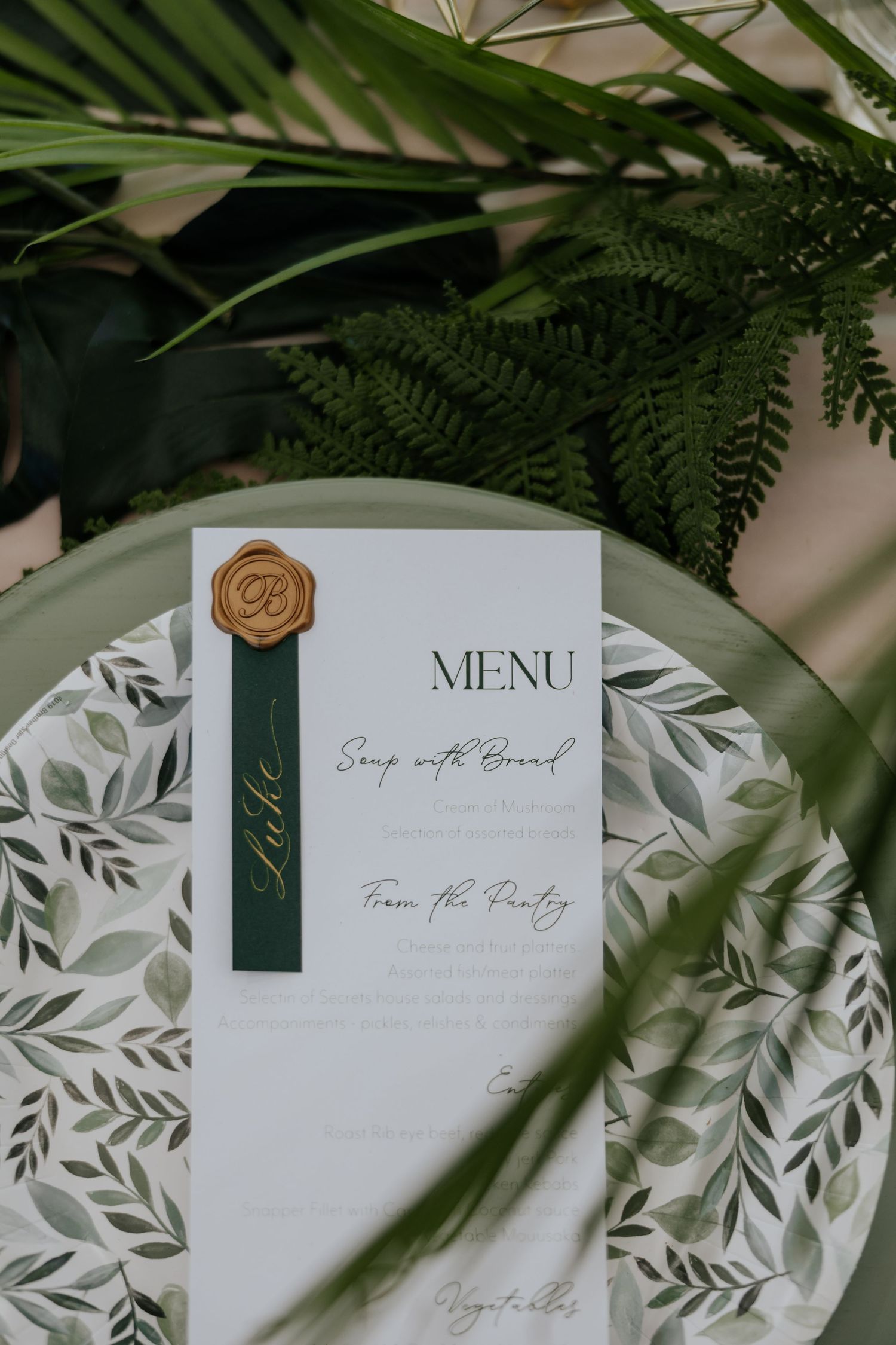 Wedding menu card with gold wax seal on botanical plate surrounded by tropical fern greenery.