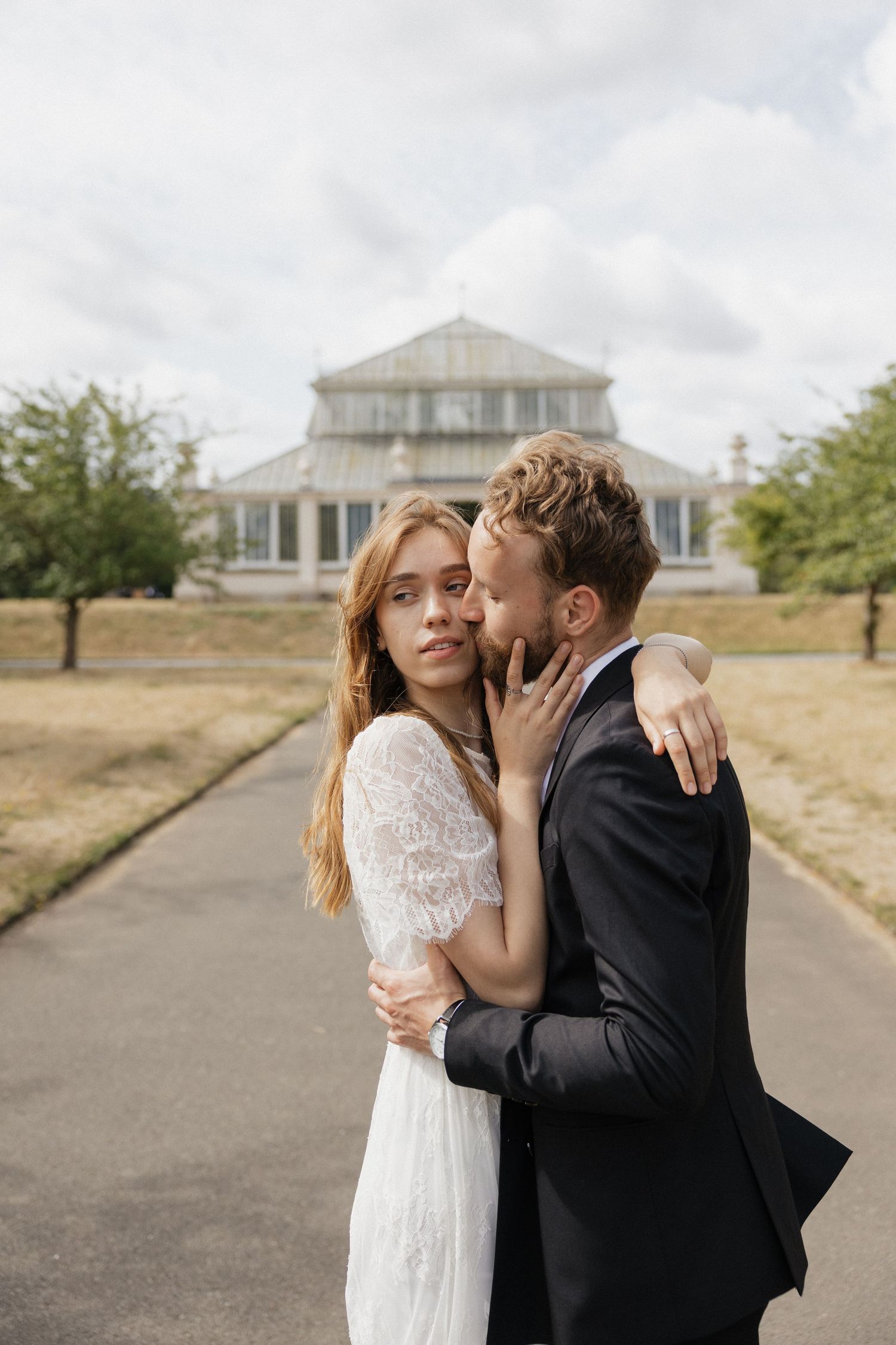 Groom whispering to bride in lace dress with a glass greenhouse building in background.