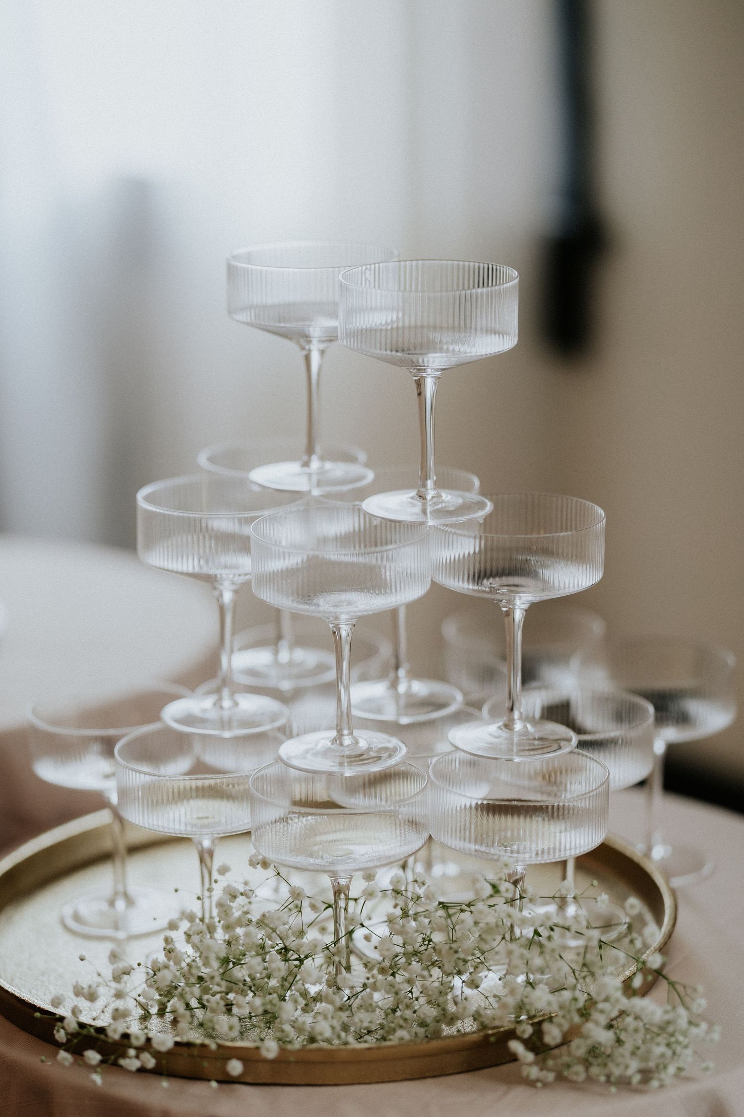 Elegant champagne tower with ribbed coupe glasses on a gold tray, decorated with delicate white baby's breath flowers.