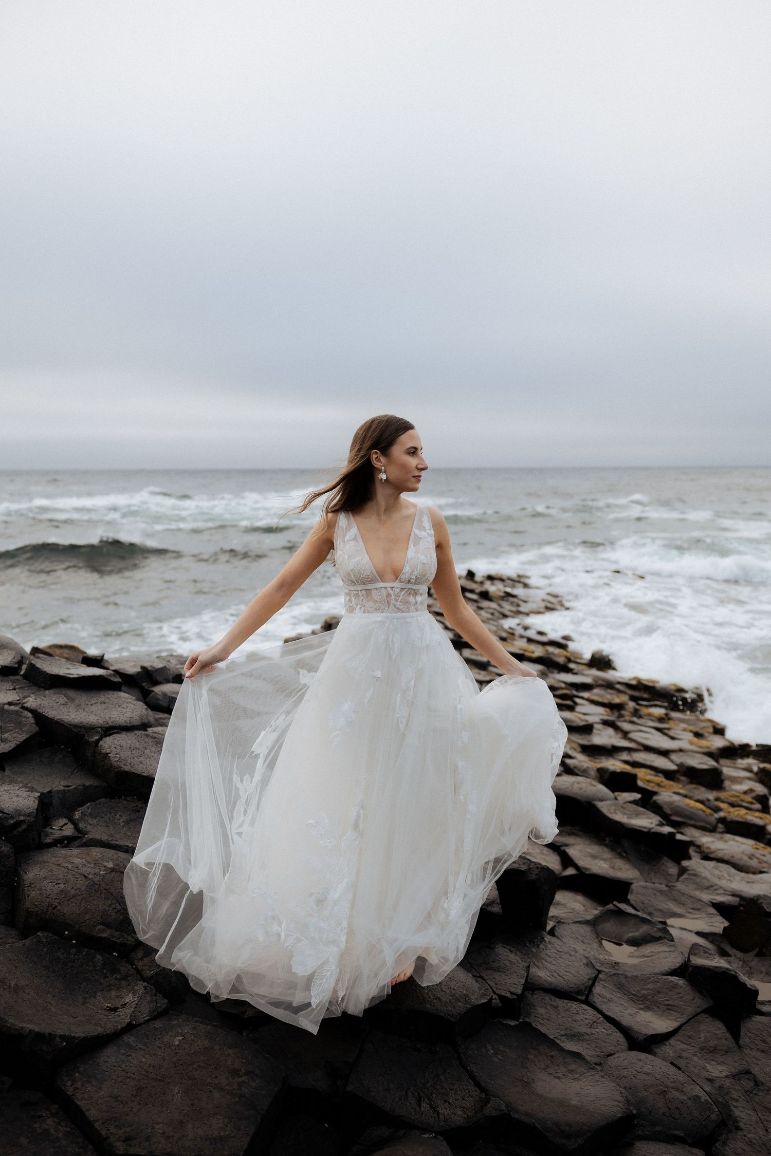 Bride in flowing white gown standing on rocky coastline with crashing ocean waves behind her.