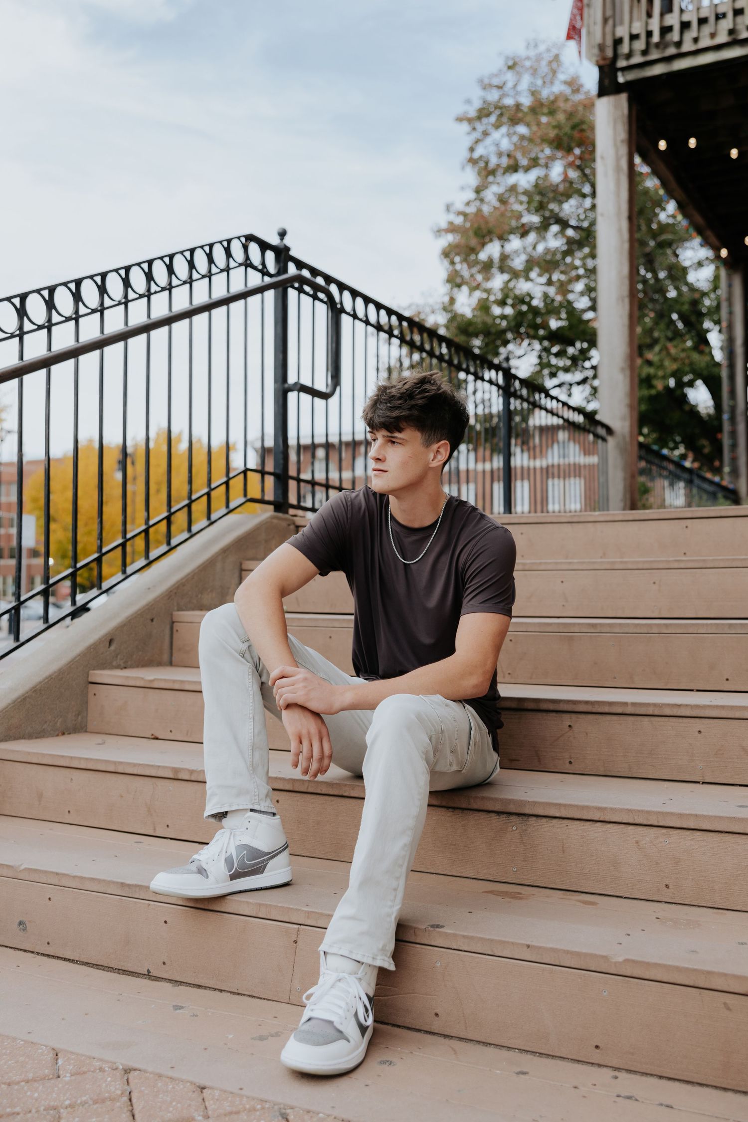 Young man in brown shirt and gray jeans sits on outdoor steps with iron railing and fall foliage.