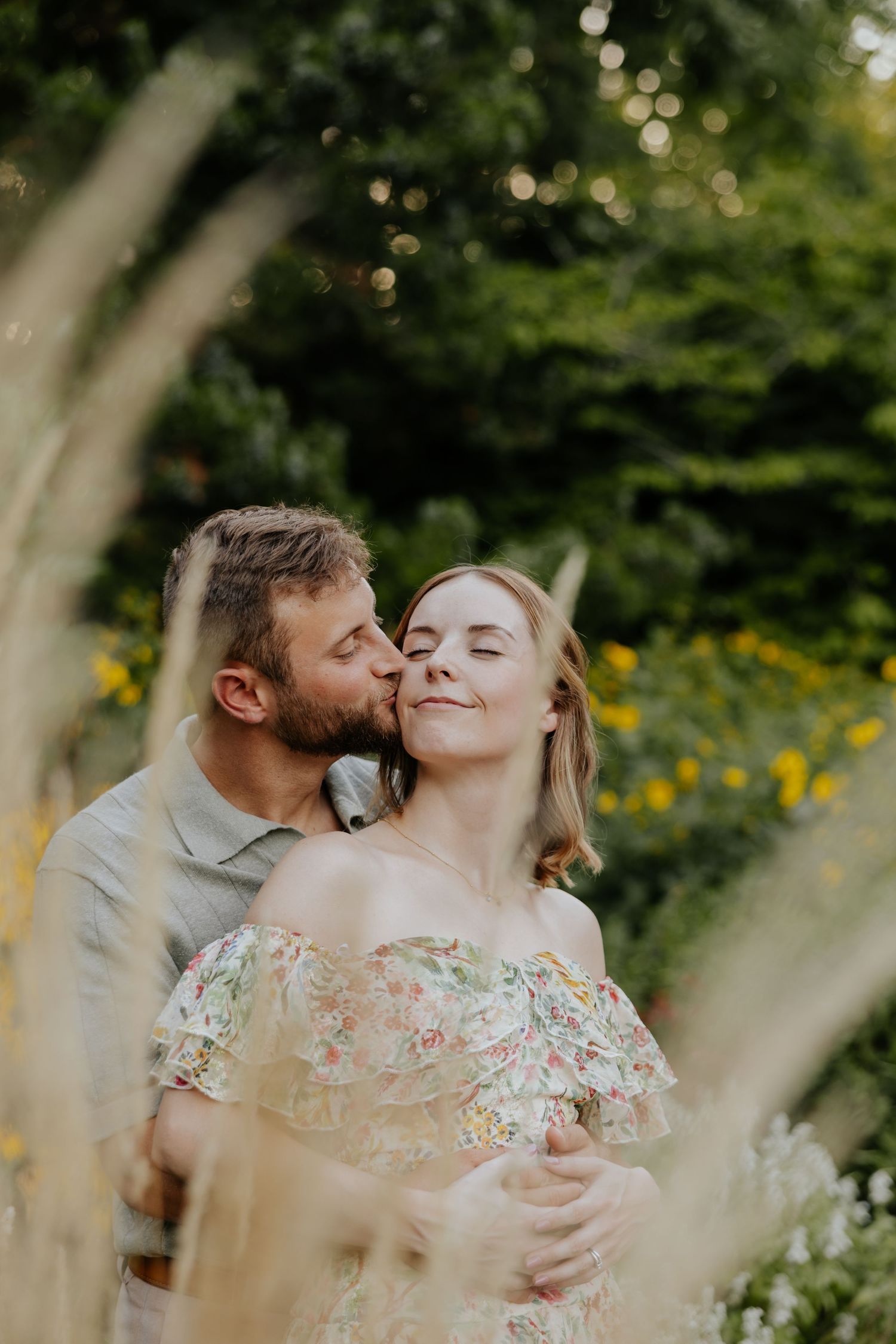 Man kisses woman on cheek as she smiles serenely in a floral dress among wildflowers.
