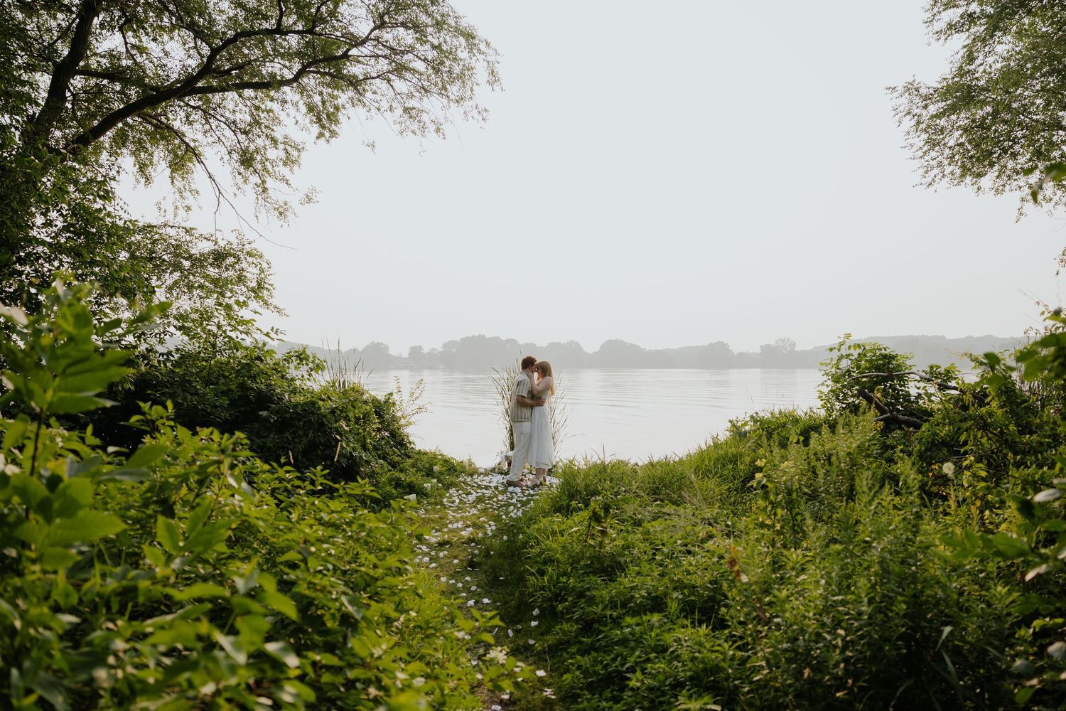 Couple embracing and kissing at a lush green lakeside landscape framed by overhanging tree branches.