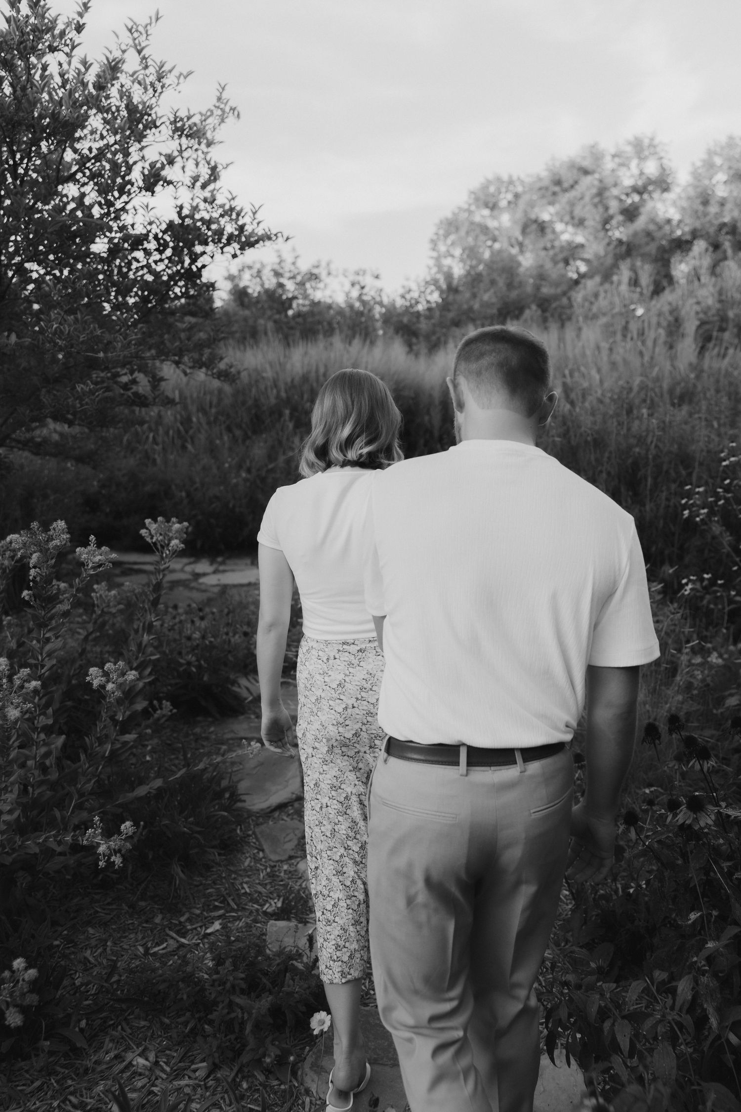 Black and white photo of a couple walking hand in hand through a lush garden path.