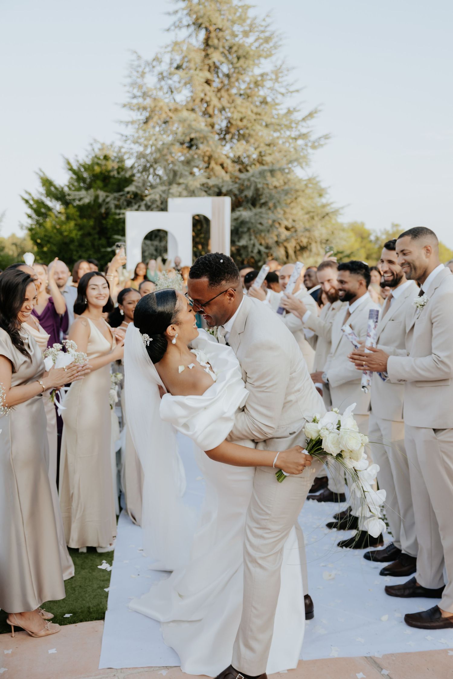 Groom lifting bride in white off-shoulder gown during joyful outdoor wedding ceremony recessional with guests celebrating.