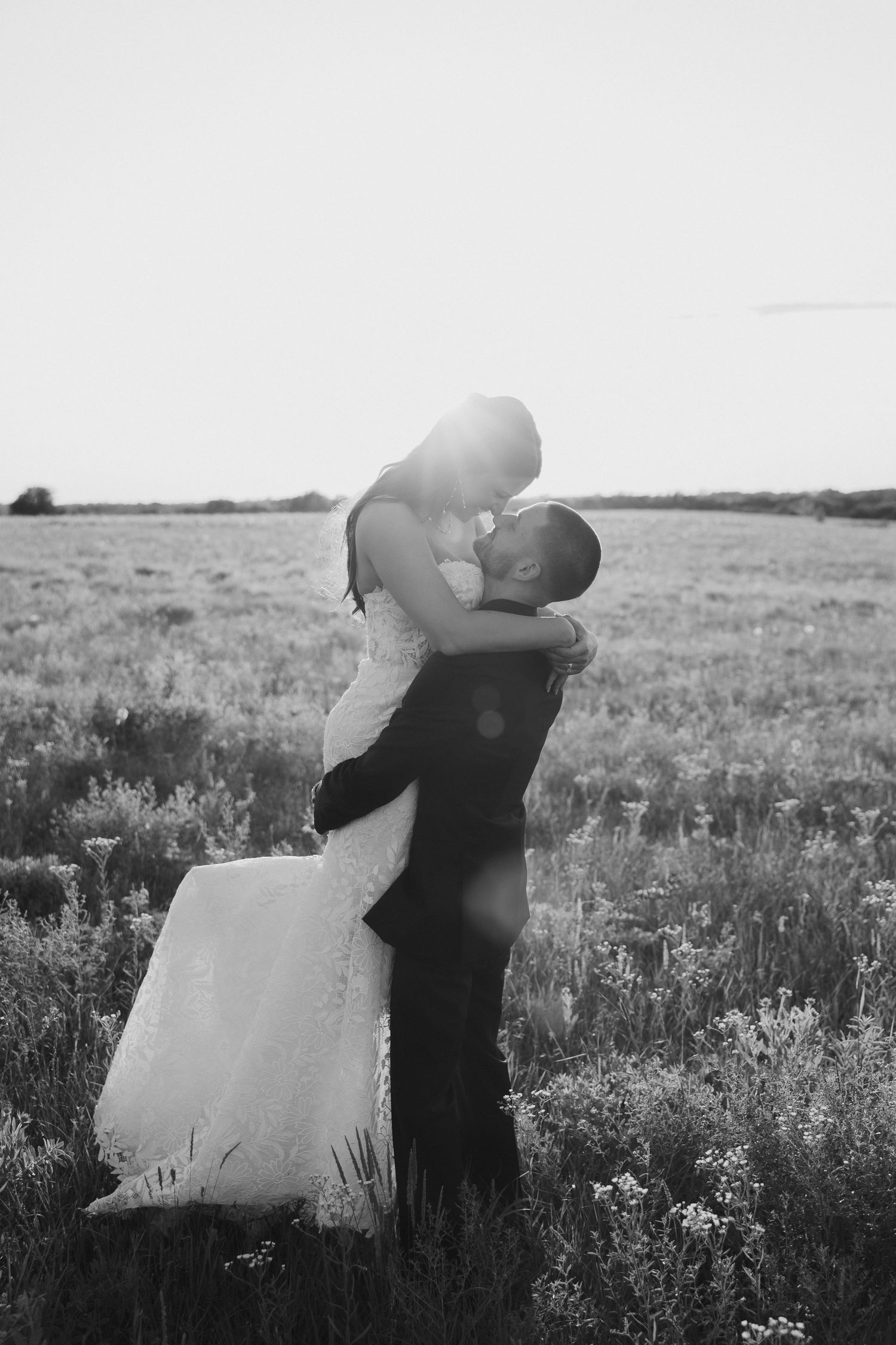 Black and white image of a groom lifting his bride in a sun-drenched meadow during their wedding day.