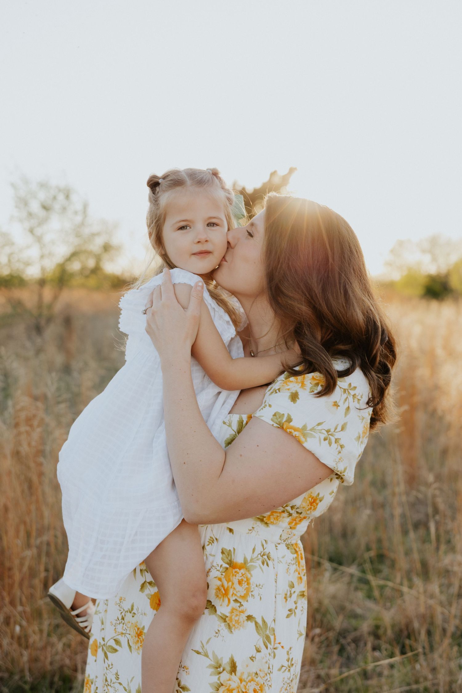 Pregnant mother kisses her young daughter in a white dress during a warm sunset outdoor portrait.