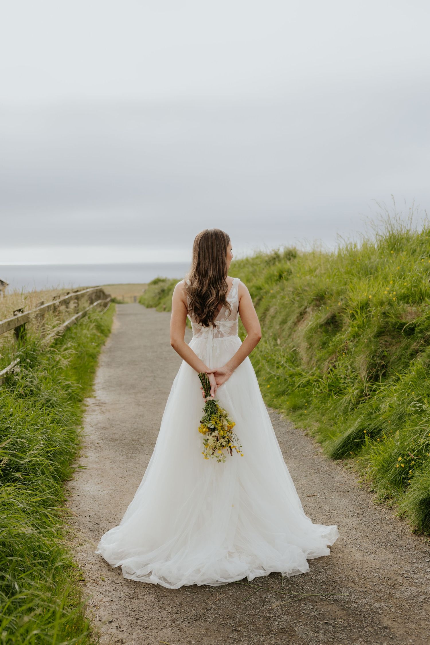 Bride in elegant white tulle gown holding yellow wildflowers on a coastal cliffside path.