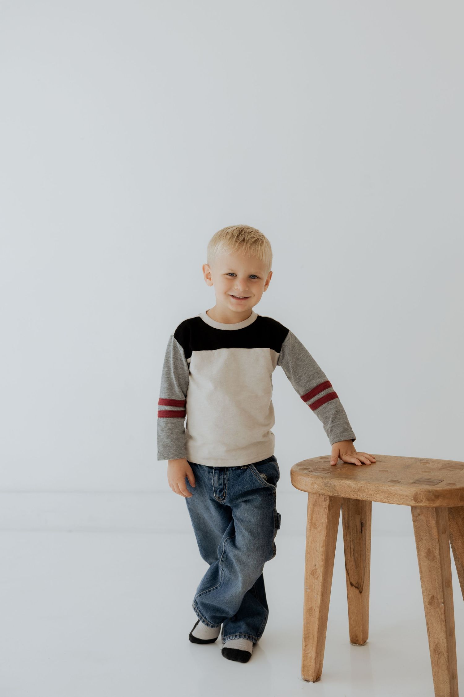 Smiling blonde boy in striped long-sleeve shirt standing next to wooden stool.