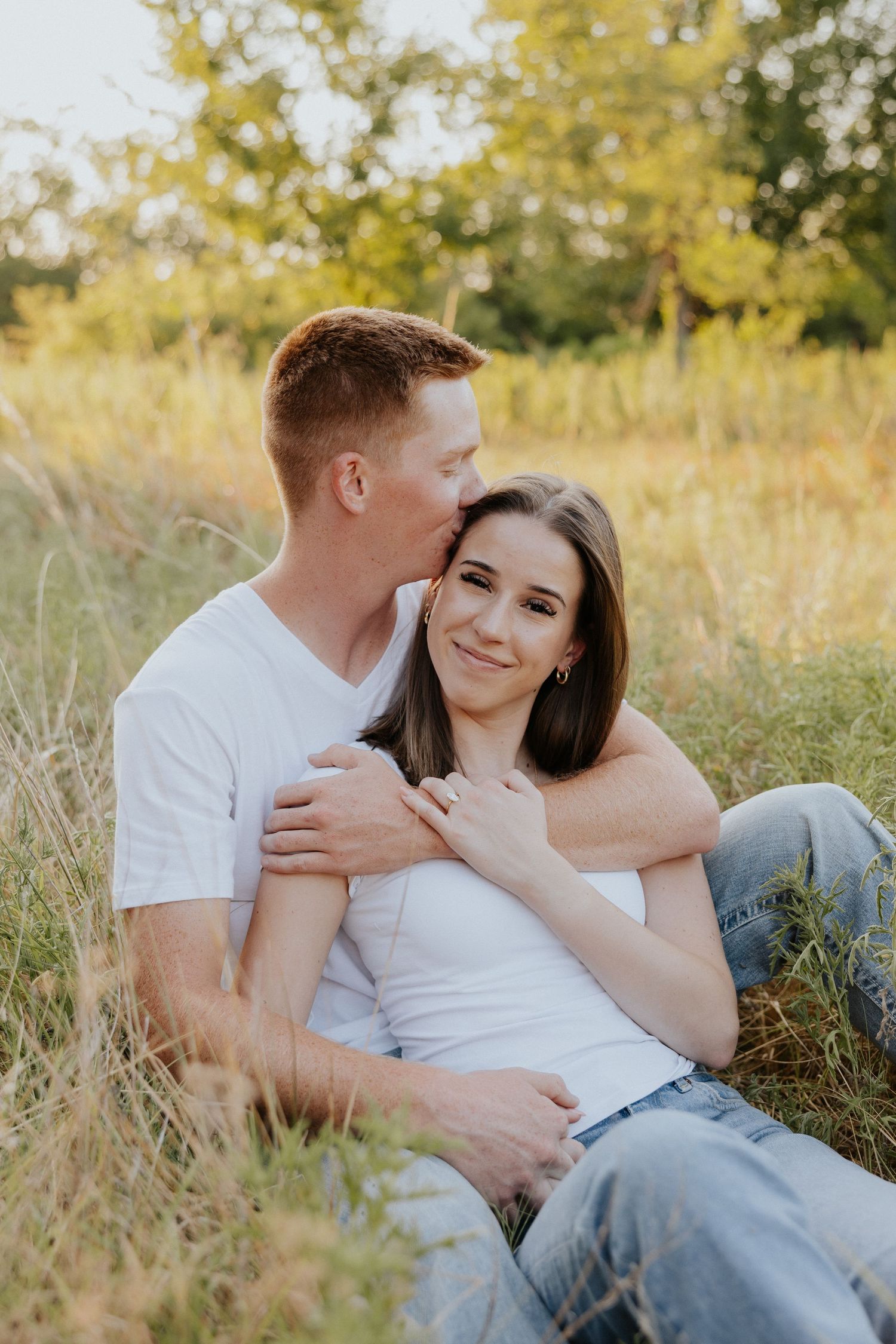 Couple sitting in golden meadow grass, man kissing woman's temple during engagement session.