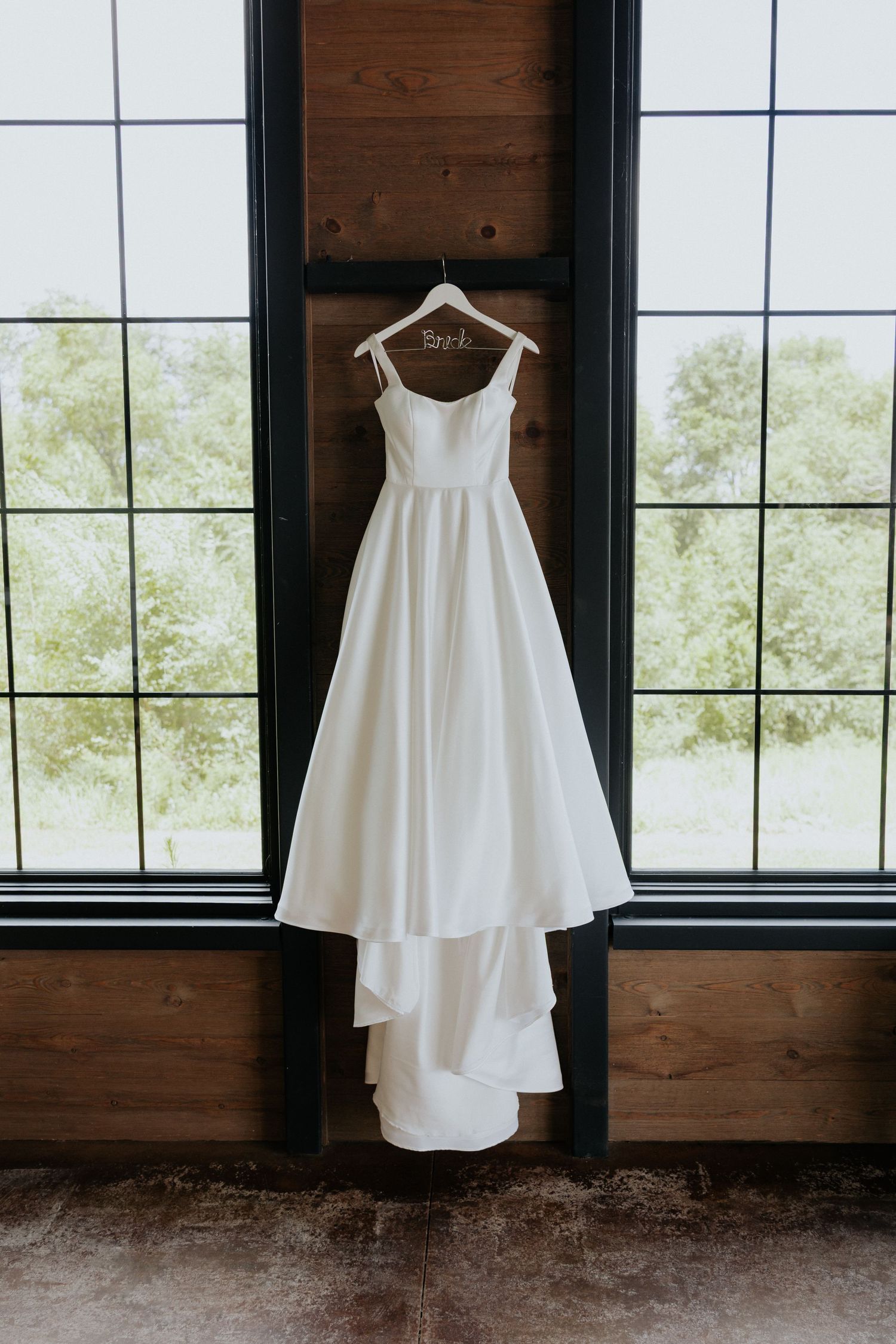Simple white A-line wedding dress hanging on personalized hanger in front of large windows.