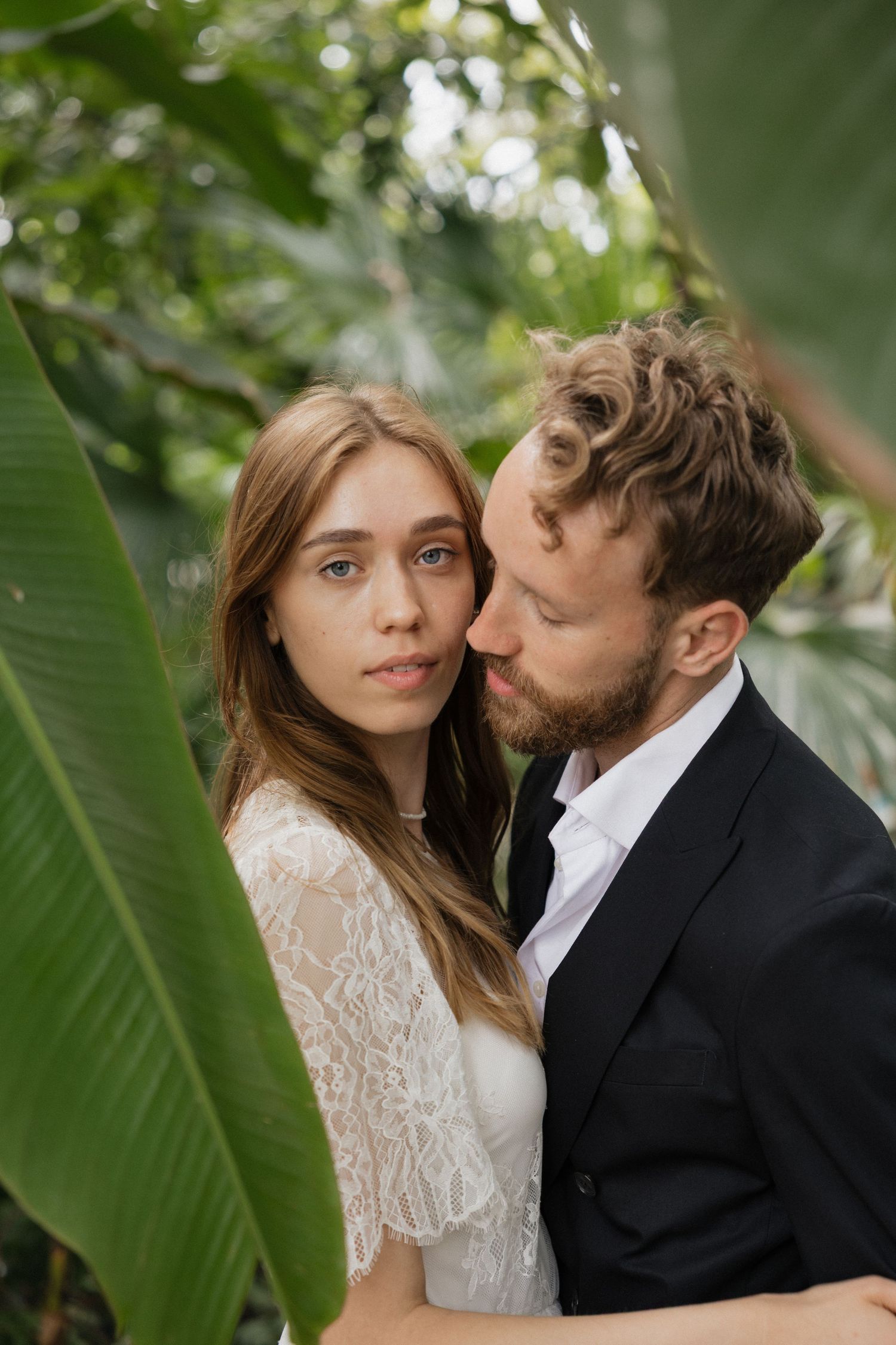 Bride in lace dress and groom in black suit embracing among large tropical green leaves.