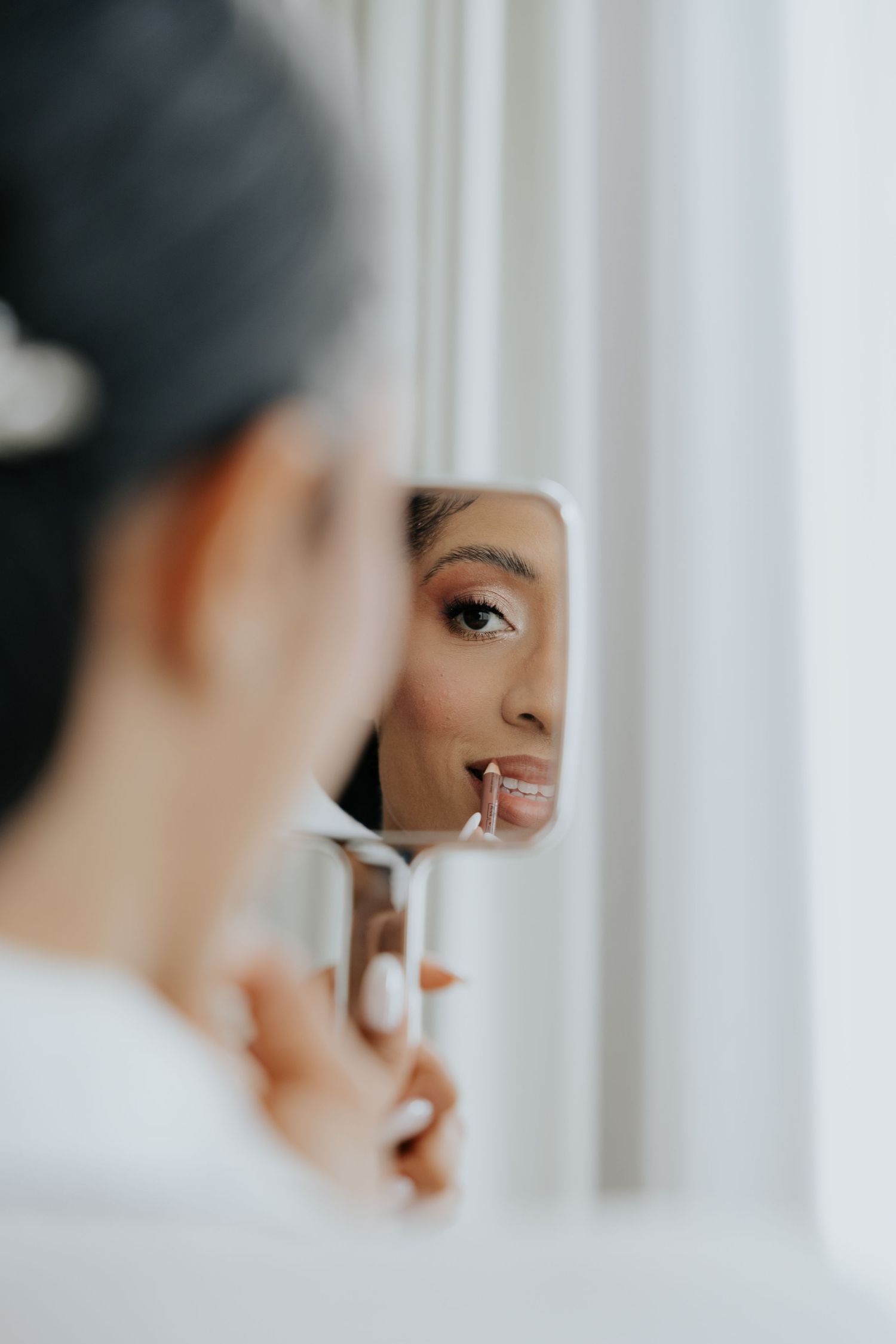 Bride applies lipstick while looking into a small handheld mirror on wedding day.