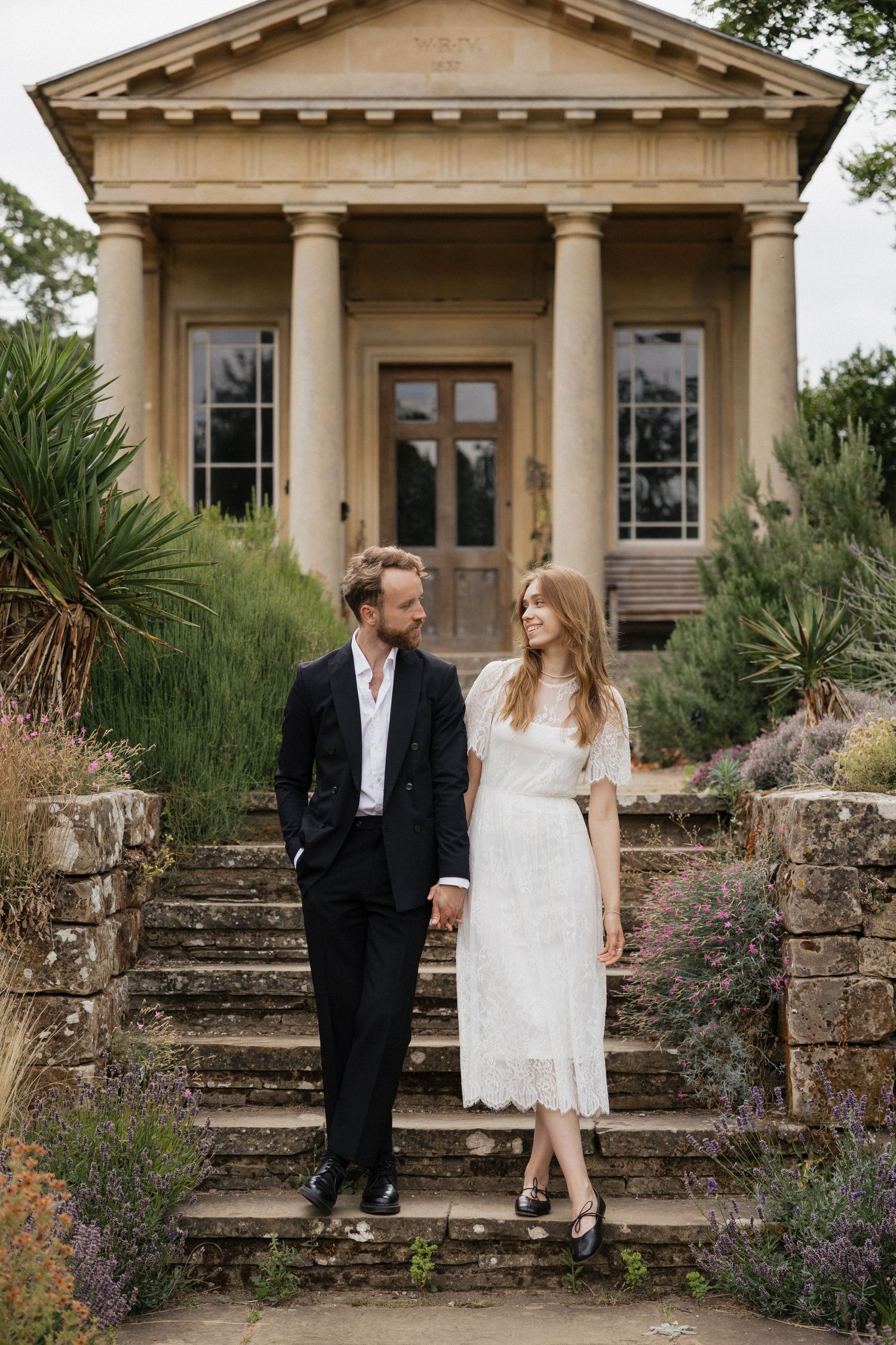 Bride and groom holding hands on stone steps before a neoclassical building surrounded by lavender gardens.