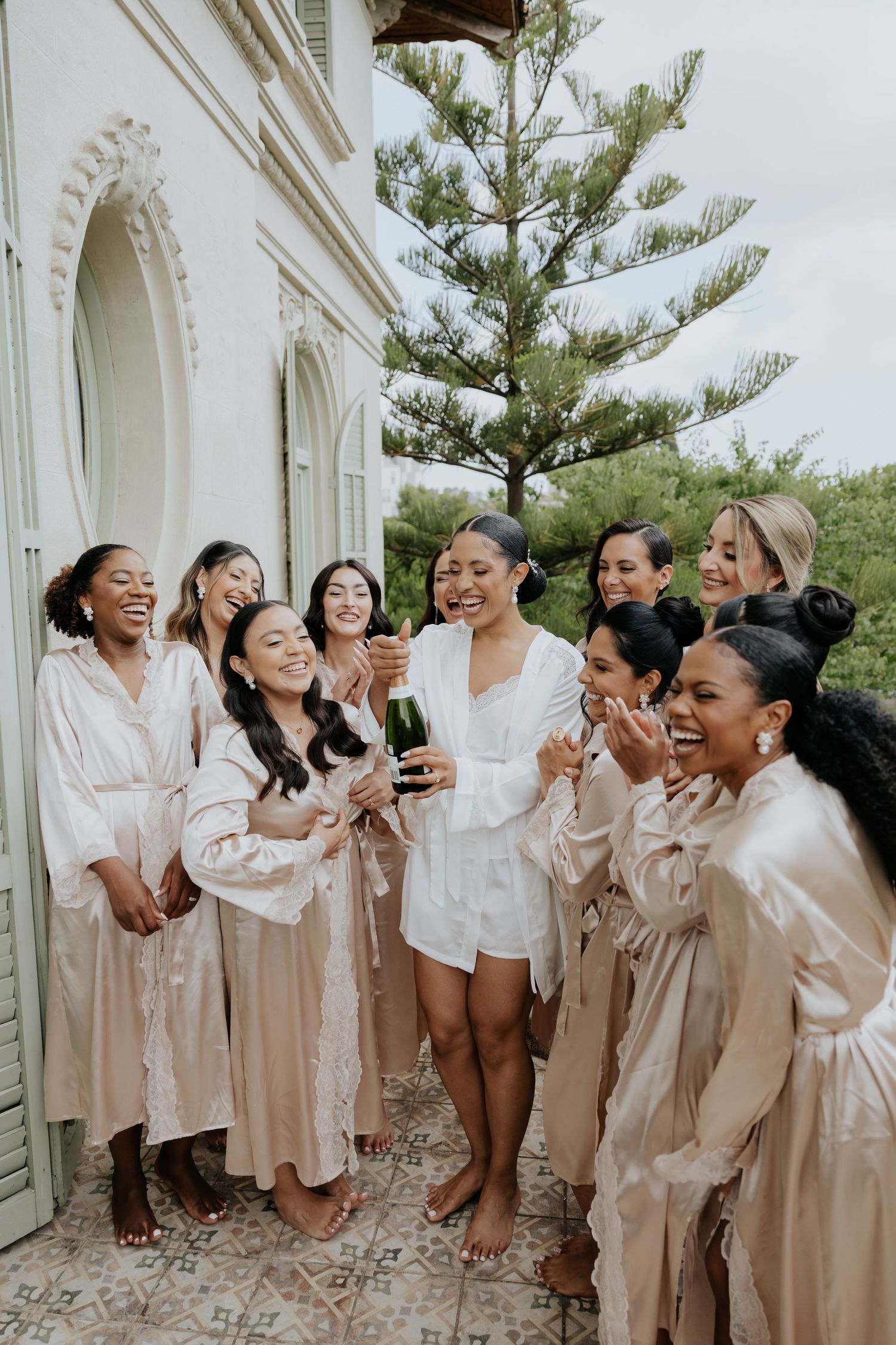 Bride in white romper pops champagne with bridesmaids in blush satin robes on a villa balcony.