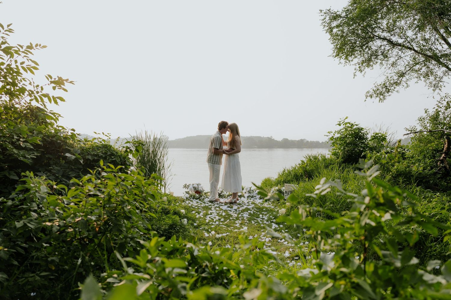Couple sharing a kiss framed by green foliage on a misty lakeside shore.