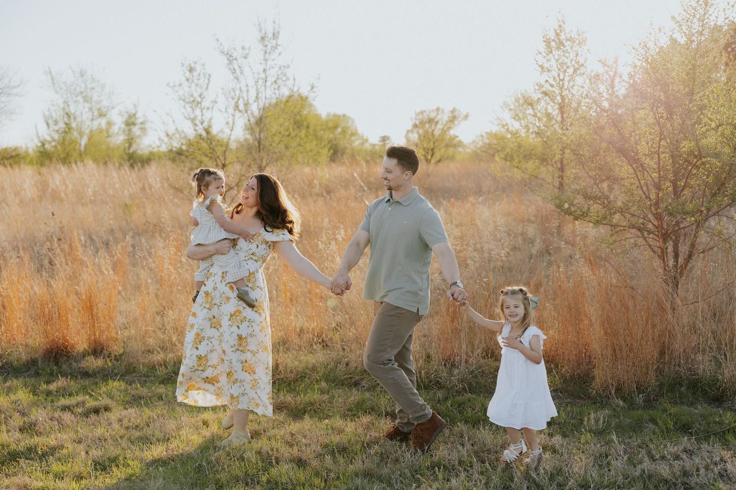 Happy family of four walks hand-in-hand through a sunlit field during a golden hour photo session.