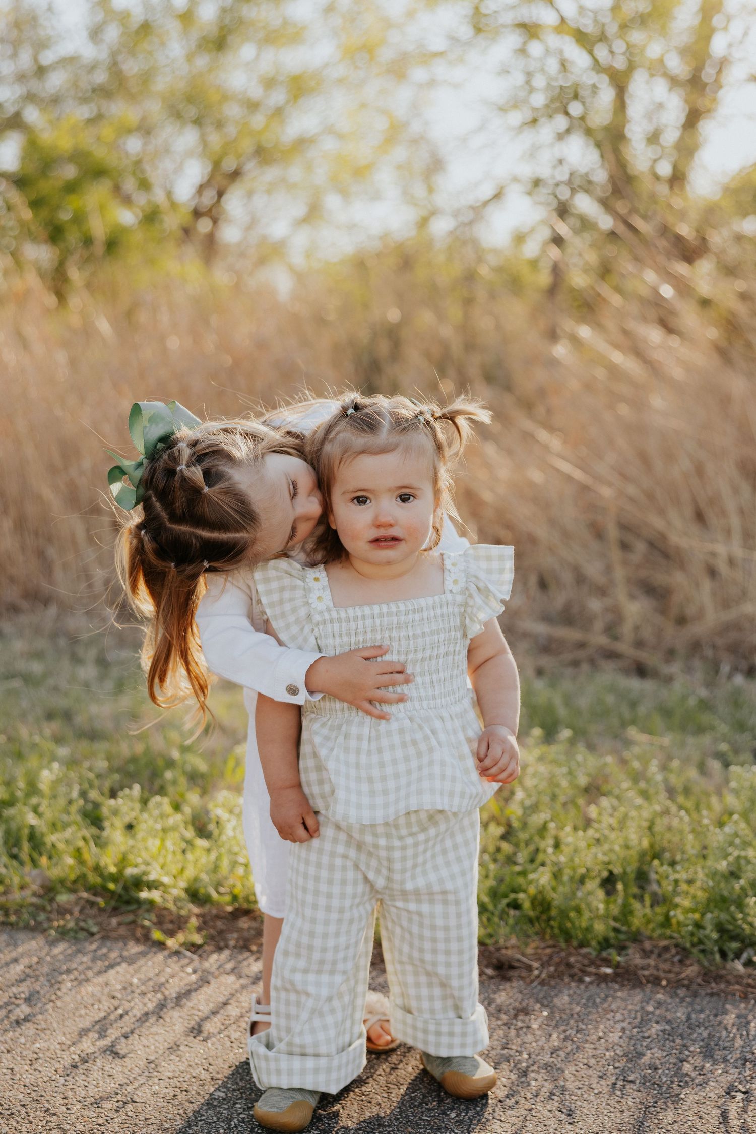 Two young sisters share a sweet moment outdoors, one kissing the other's cheek in golden hour light.