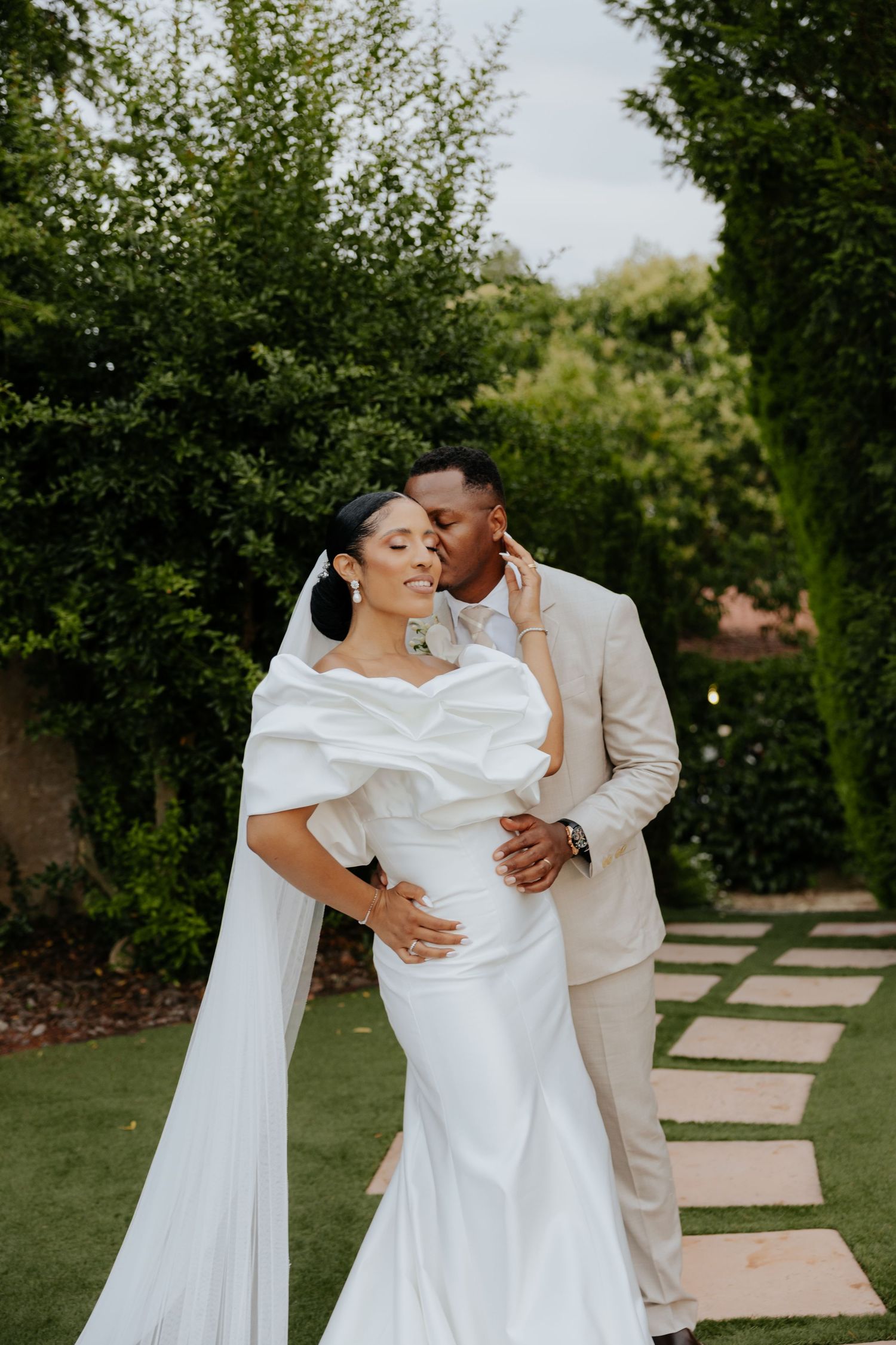 Bride in off-shoulder gown and groom in beige suit share a tender moment in a garden.