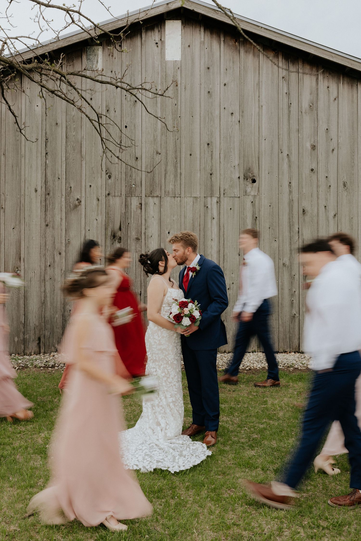 A bride and groom share a kiss outside a rustic barn while wedding guests walk past in motion blur.