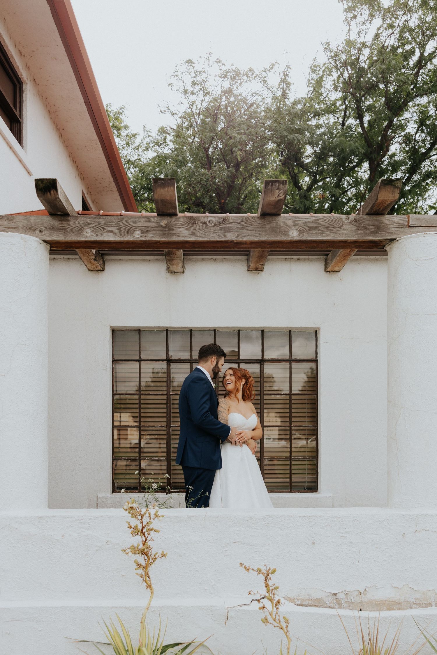 A bride and groom share a romantic moment in front of a white stucco building with wooden beams and large windows.