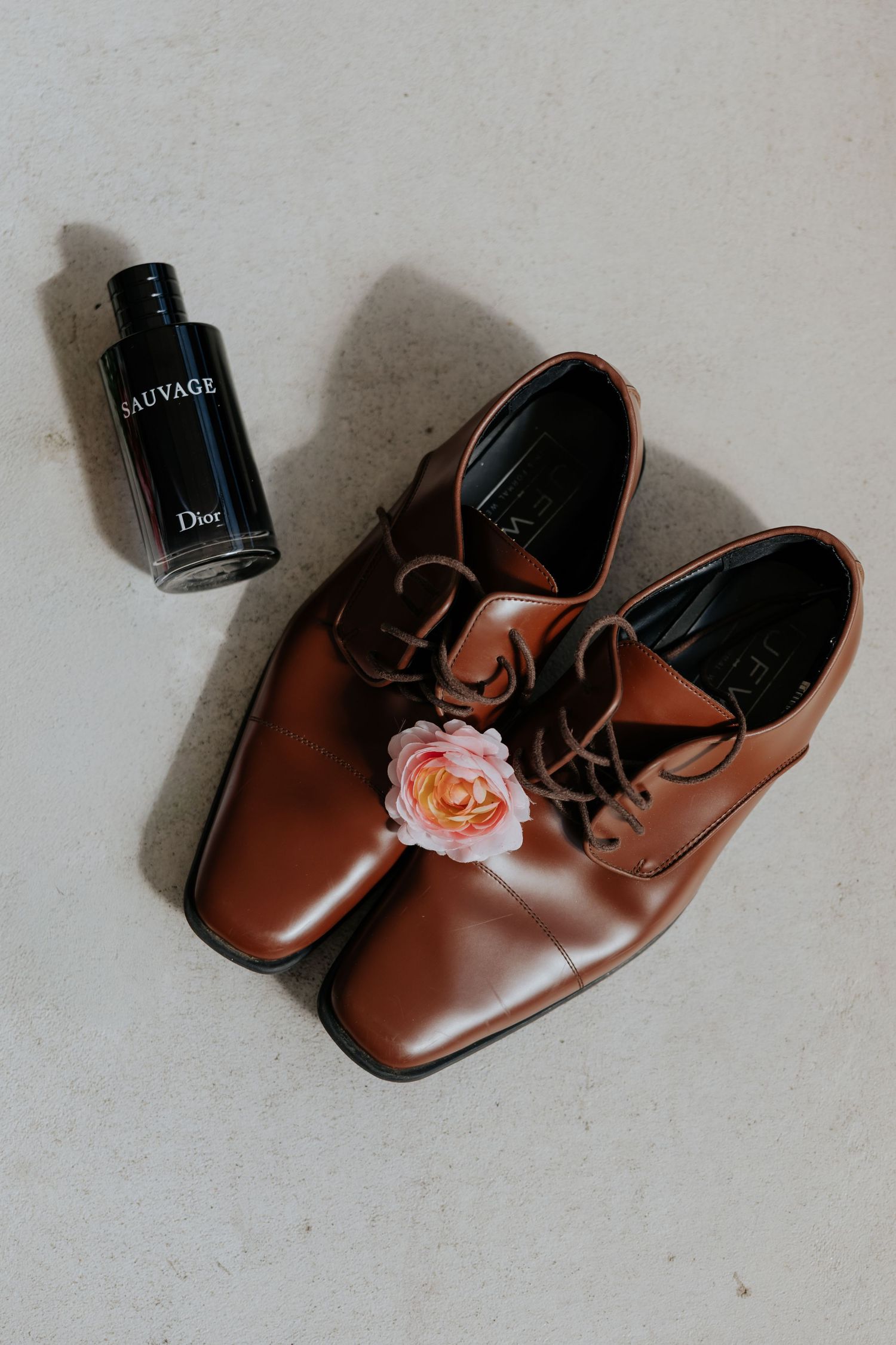 Brown leather dress shoes with pink rose boutonniere beside Dior Sauvage cologne bottle.
