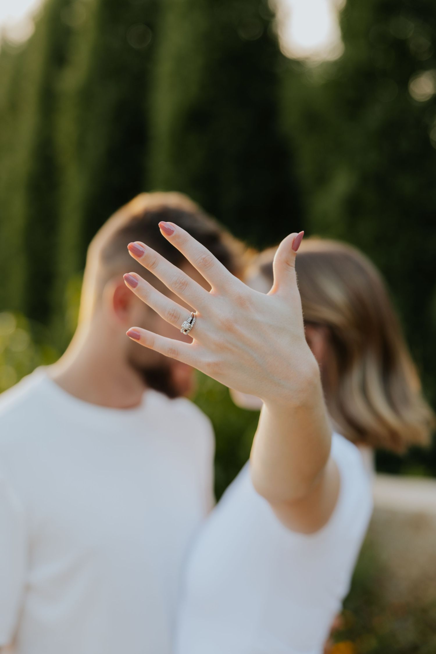 A woman shows off her engagement ring to the camera while kissing her partner outdoors near cypress trees.