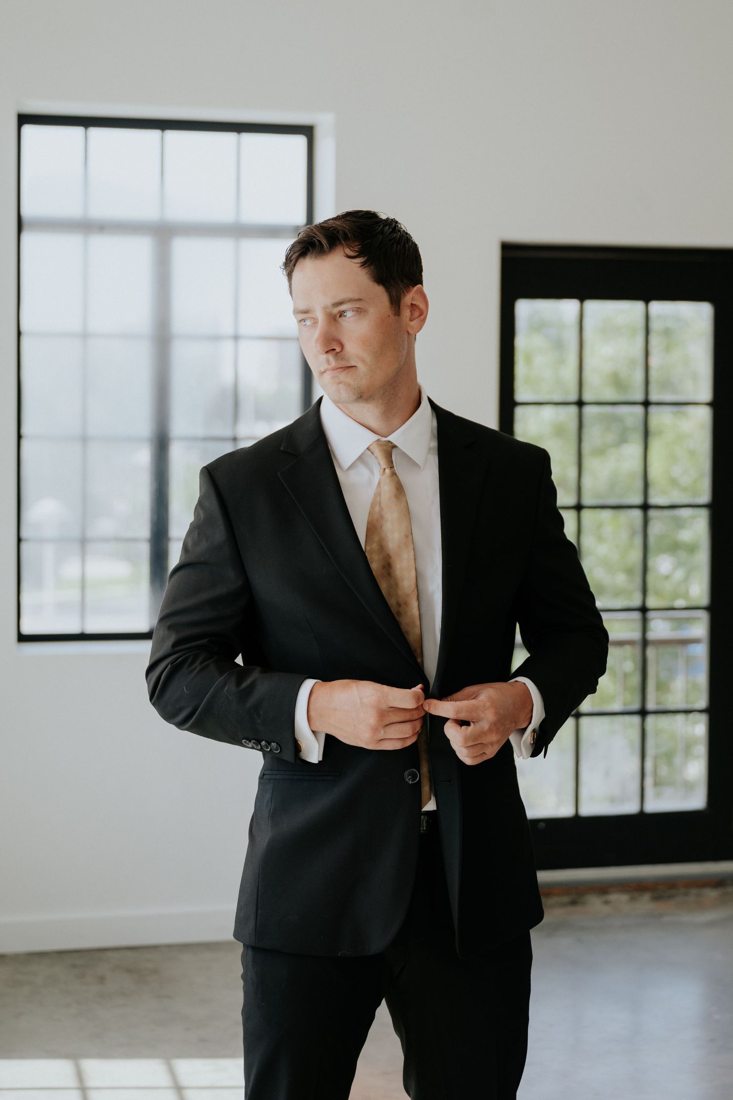 Groom in black suit with gold tie buttoning jacket near large windows indoors.