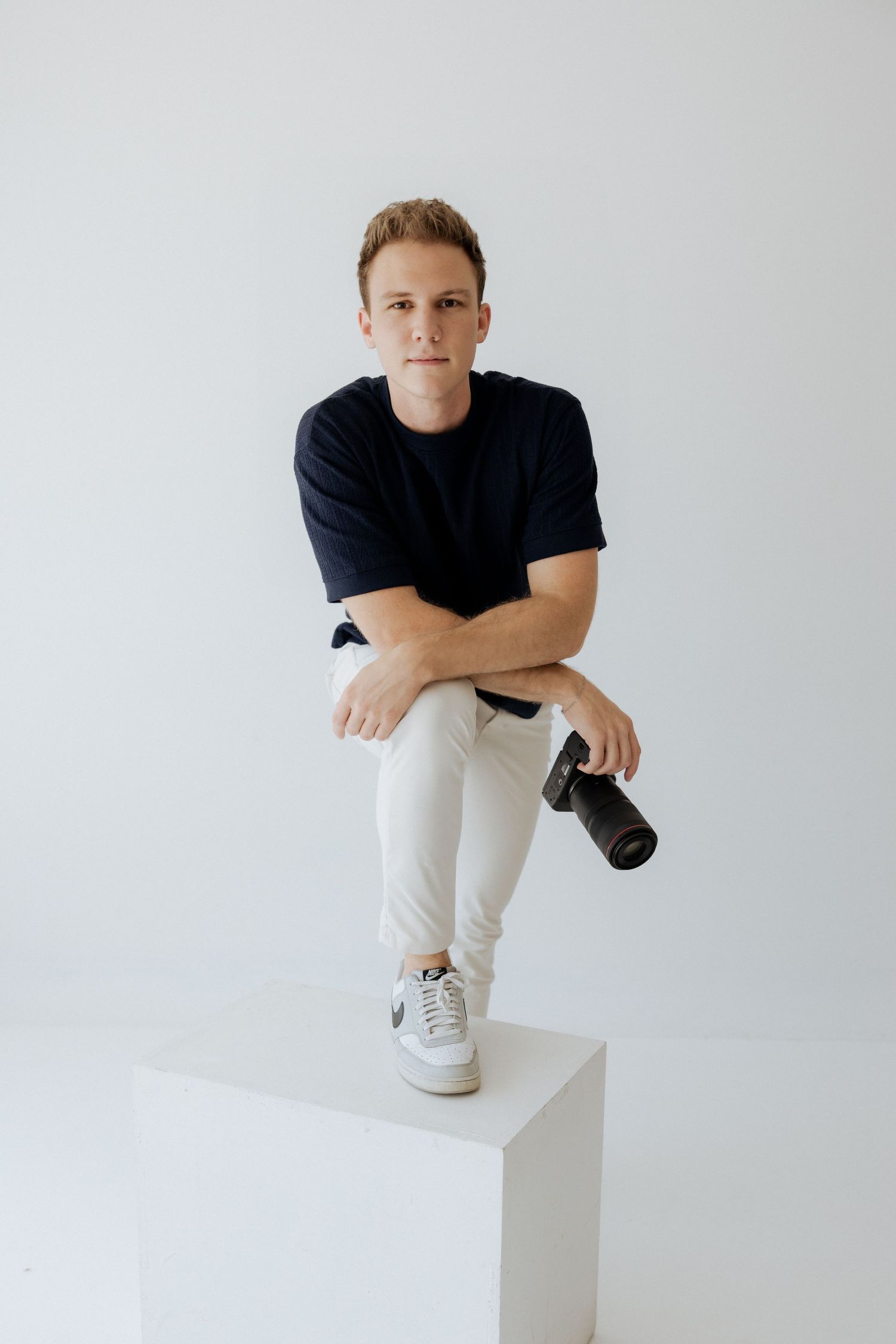 Young male photographer sitting on white cube holding camera against clean white studio background.