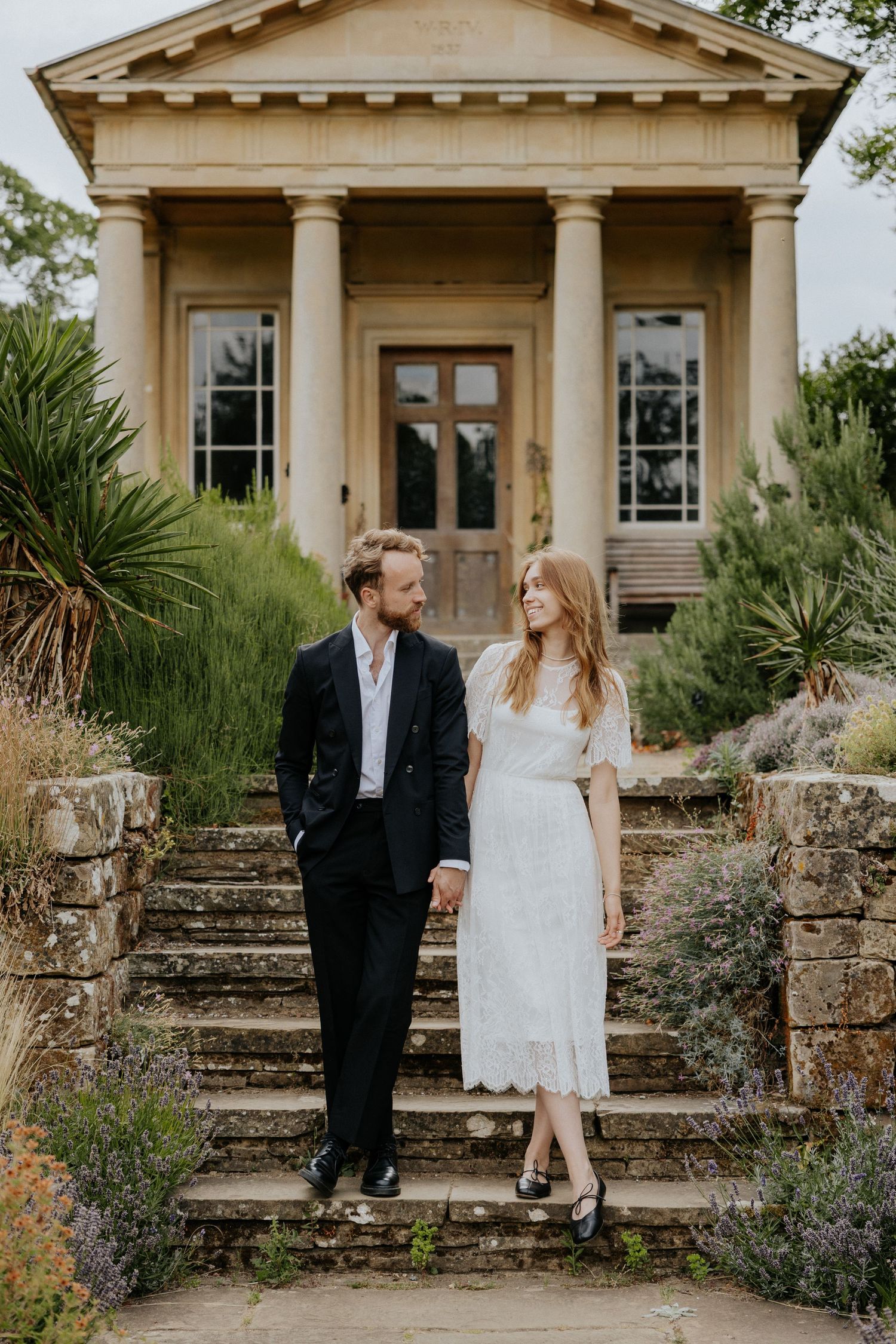 Bride and groom holding hands on stone steps before a classical temple building in a garden.