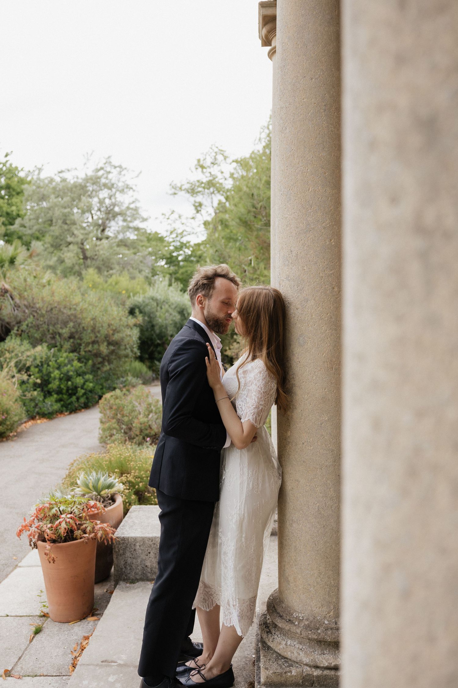 Bride and groom sharing an intimate moment leaning against a stone column in a garden setting.