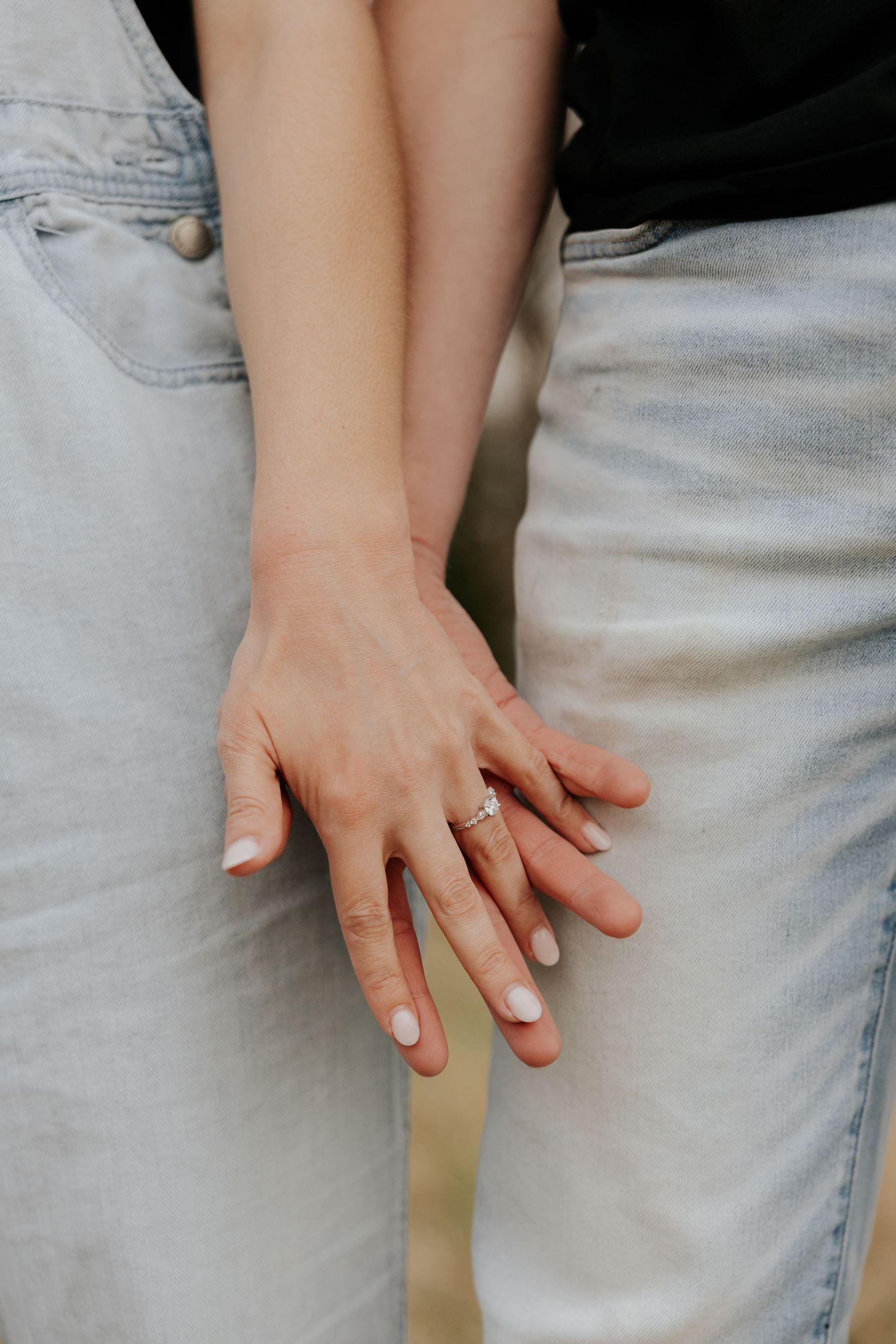 Close-up of engaged couple holding hands showing diamond engagement ring while wearing light denim outfits.