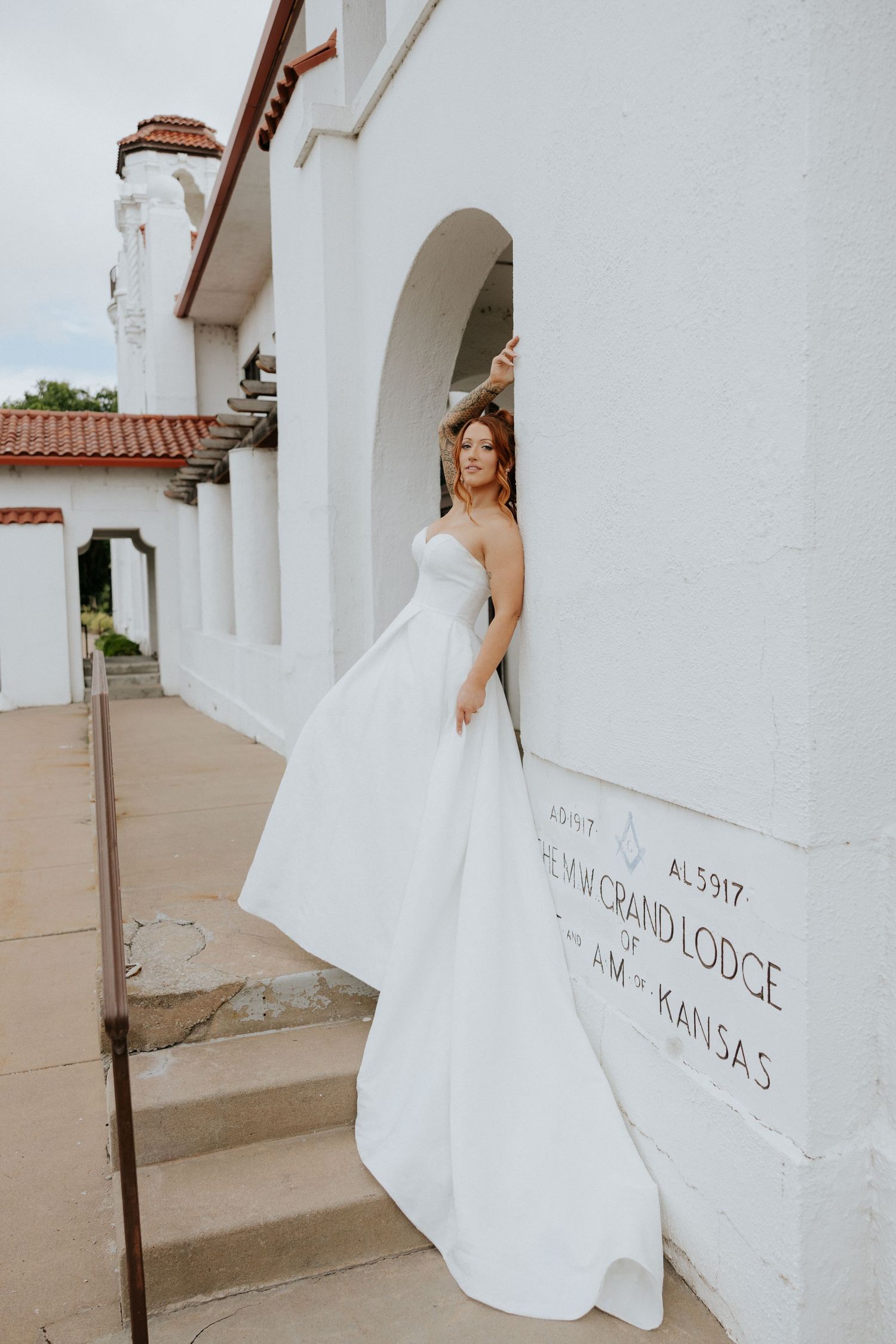 Bride in a strapless white ballgown posing at the Grand Lodge in Arkansas, Kansas, leaning against a white stucco building.