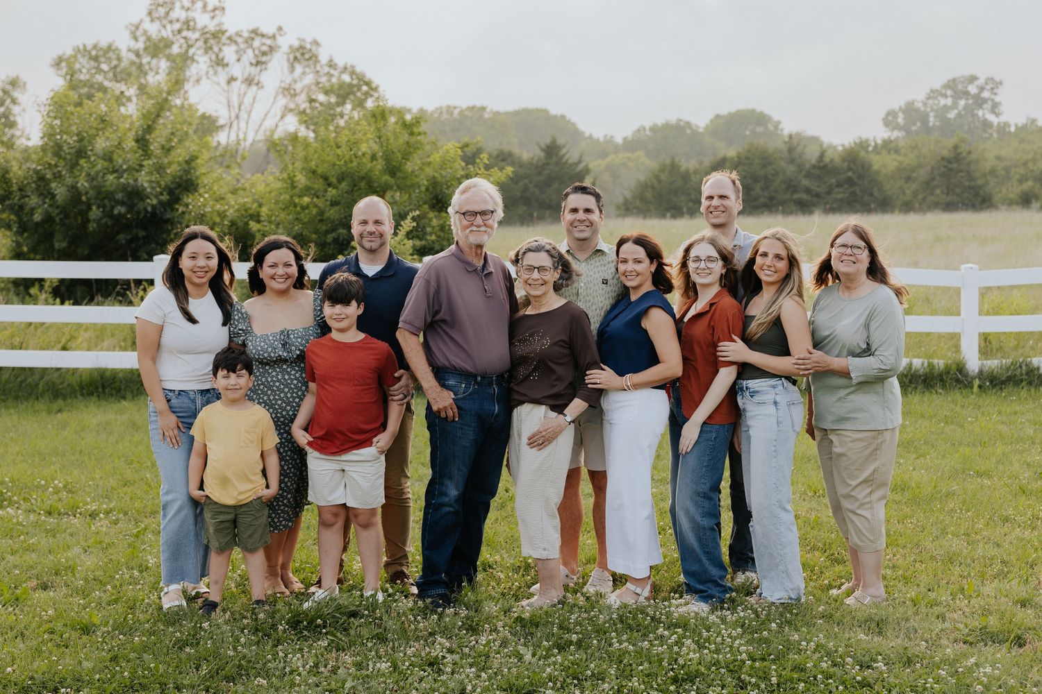Large multigenerational family poses together smiling in a green field beside a white fence.
