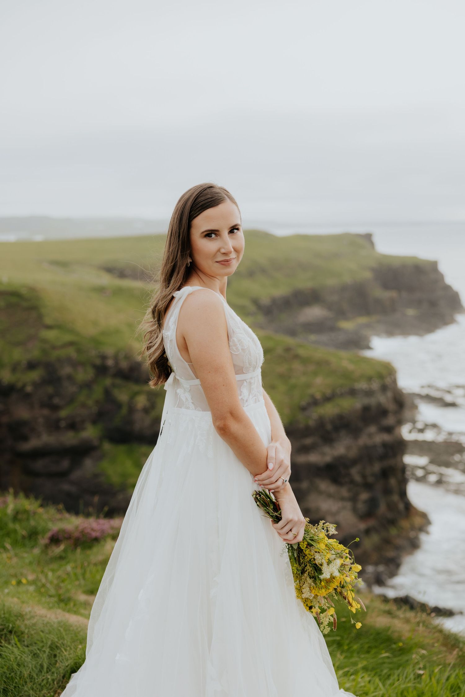 Bride in white lace gown holding yellow flowers while posing on a scenic coastal cliff.