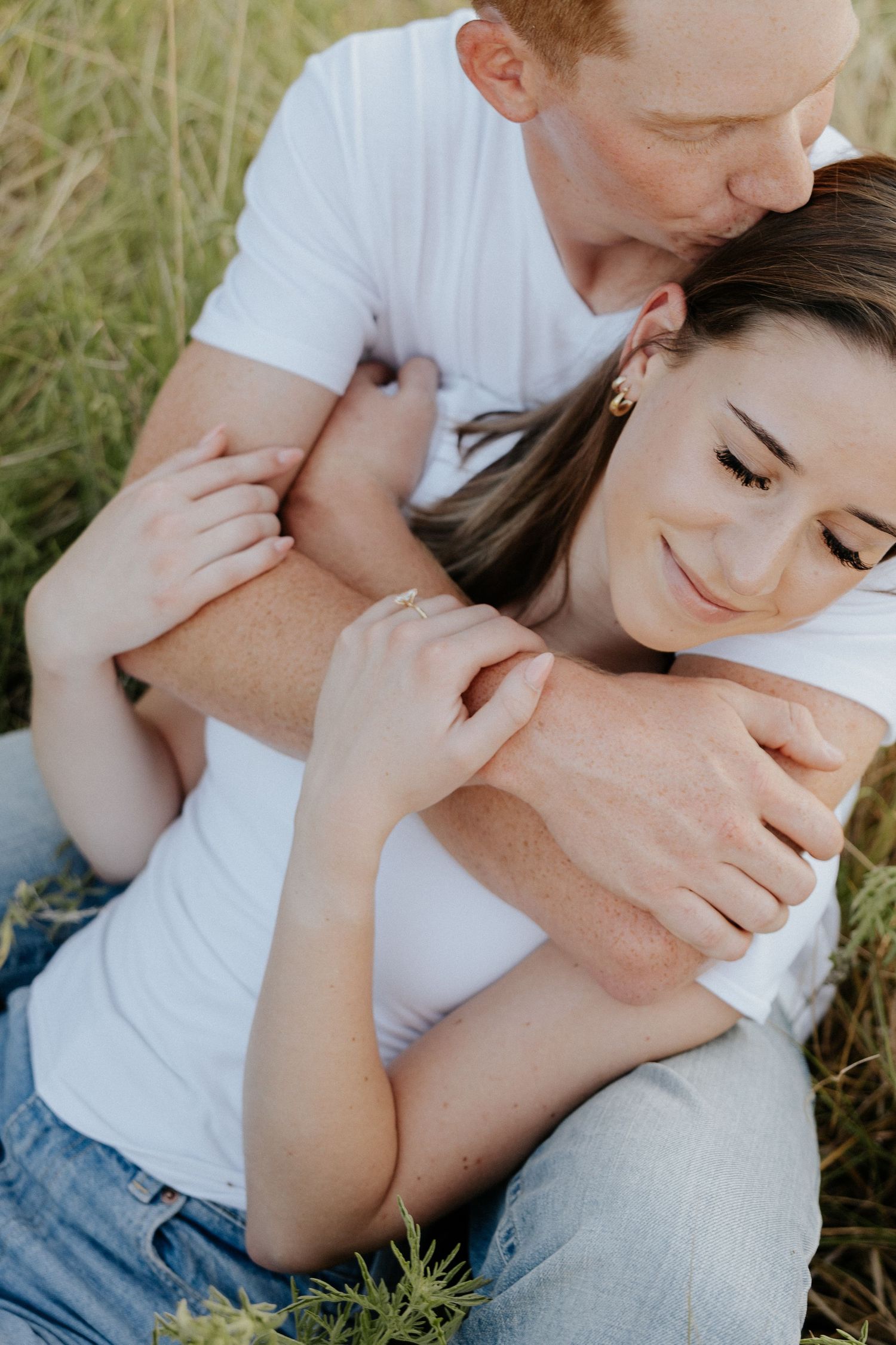 Romantic close-up of couple embracing in tall grass, woman smiling as partner kisses her head.