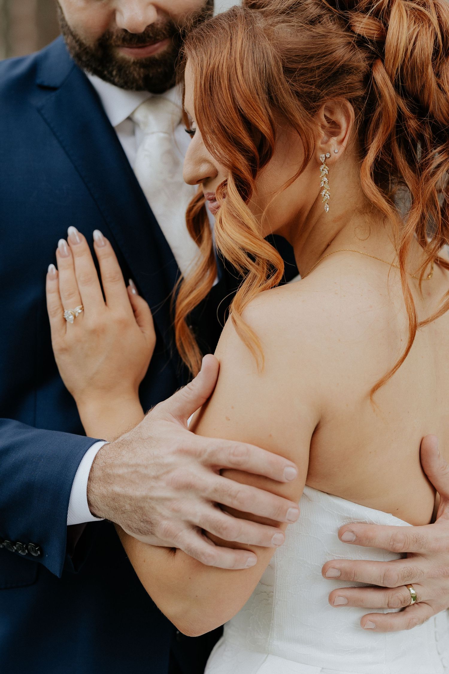 Bride and groom embrace at wedding, showing diamond engagement ring and gold wedding bands, bride in white gown with red hair.