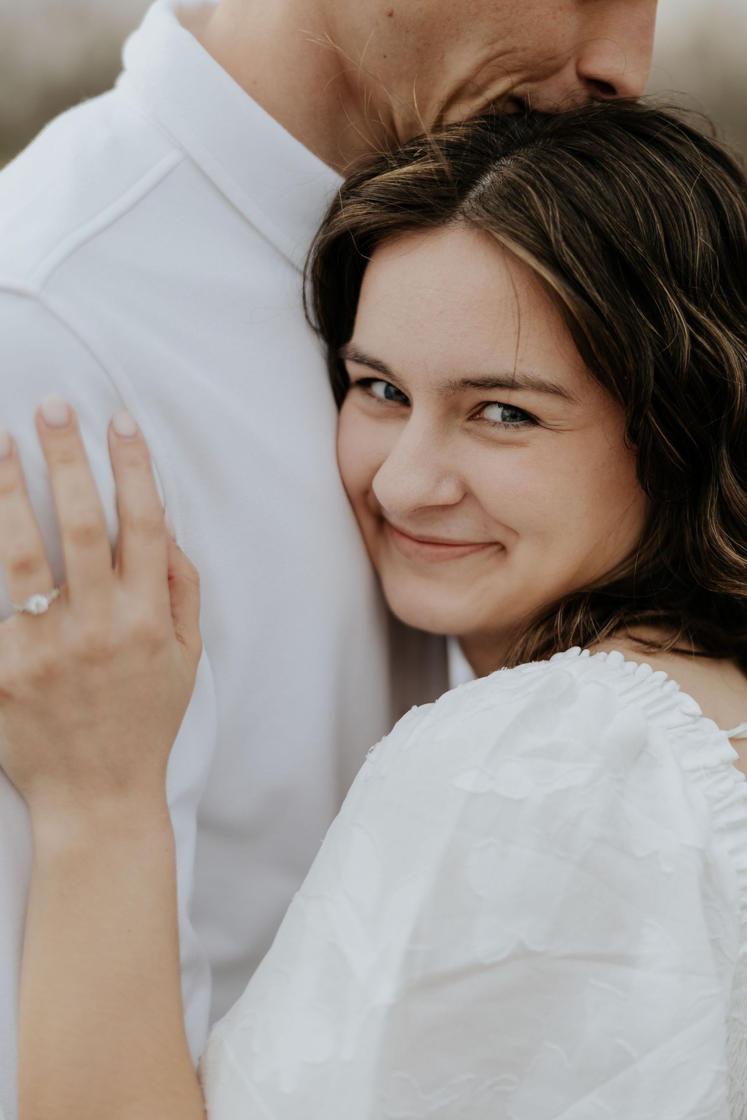 Smiling brunette woman in white dress leans against partner showing off engagement ring.